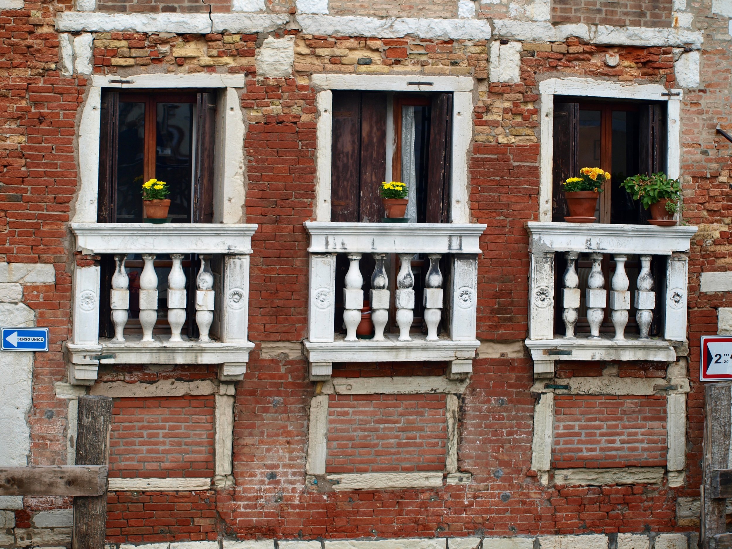 Façade d'un bâtiment avec trois fenêtres en pierre et un mur en briques rouges, quatre pots de fleurs avec des fleurs jaunes sous chaque fenêtre, un panneau de signalisation bleu et blanc à gauche et un panneau de limite de vitesse à droite.