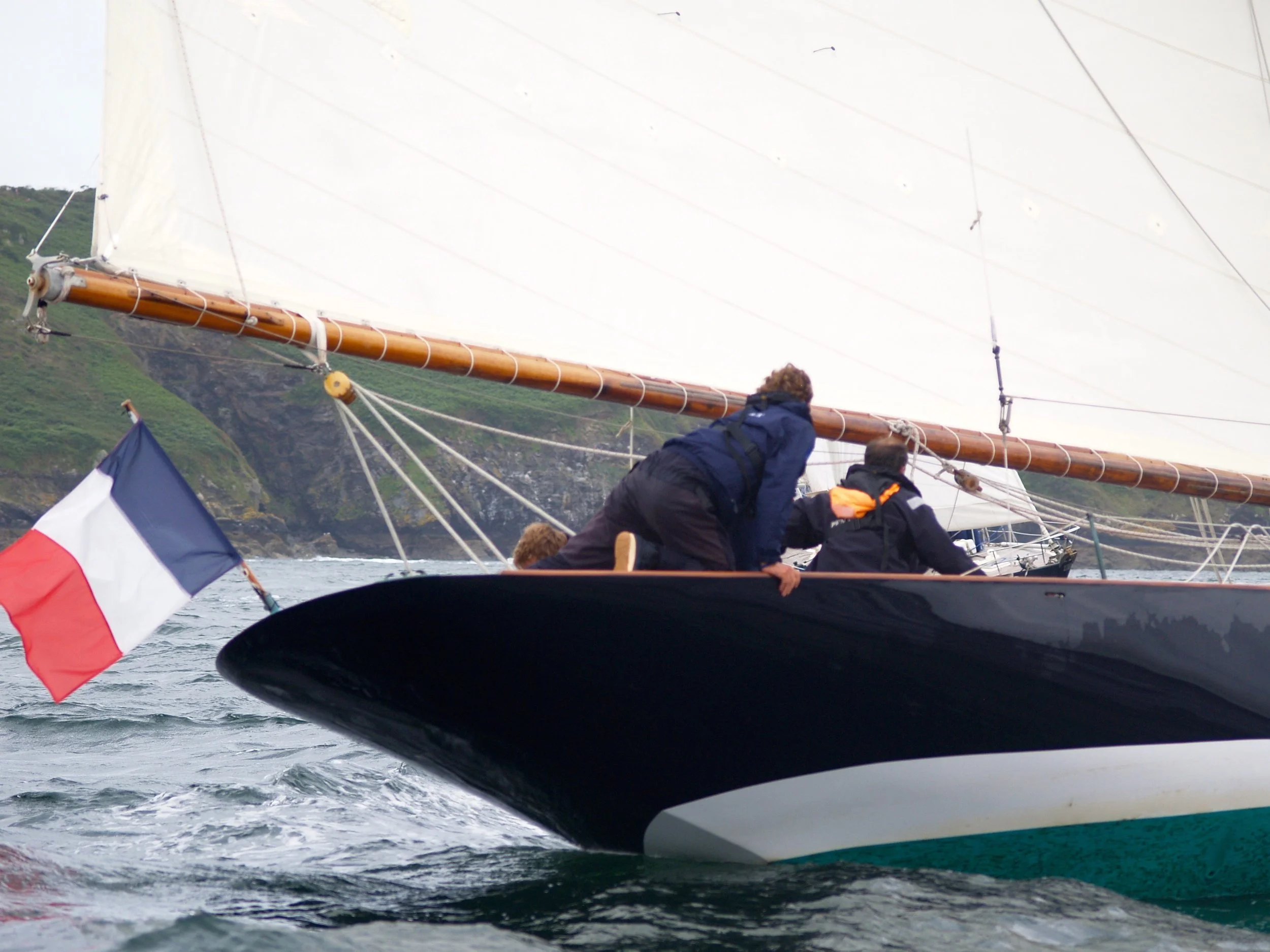 Gens en voile sur un bateau en mer, avec un drapeau français, avec un paysage de falaises en arrière-plan.