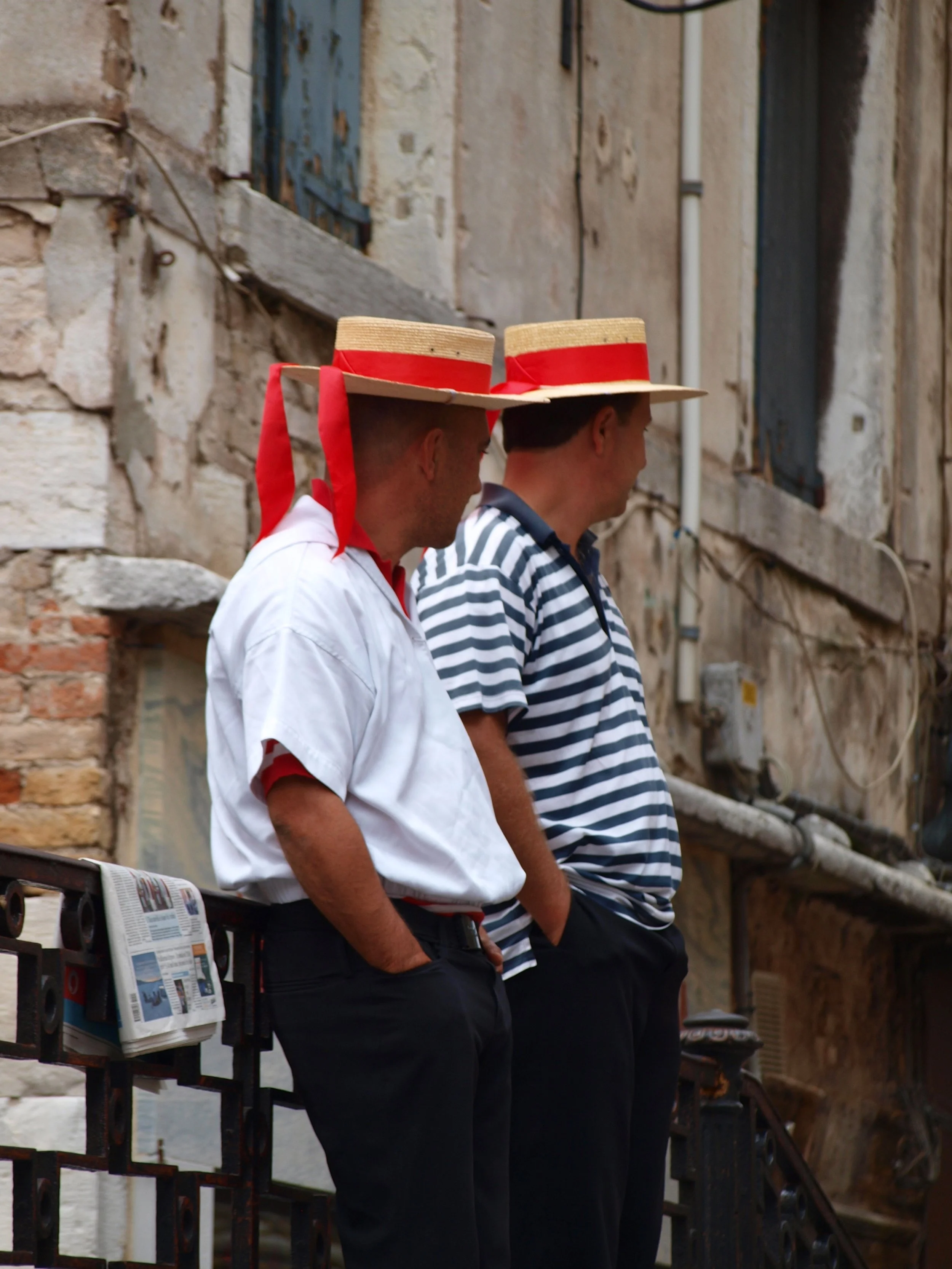 Deux hommes portant des chapeaux en paille avec un ruban rouge, debout contre un mur en pierre, regardant vers la droite.