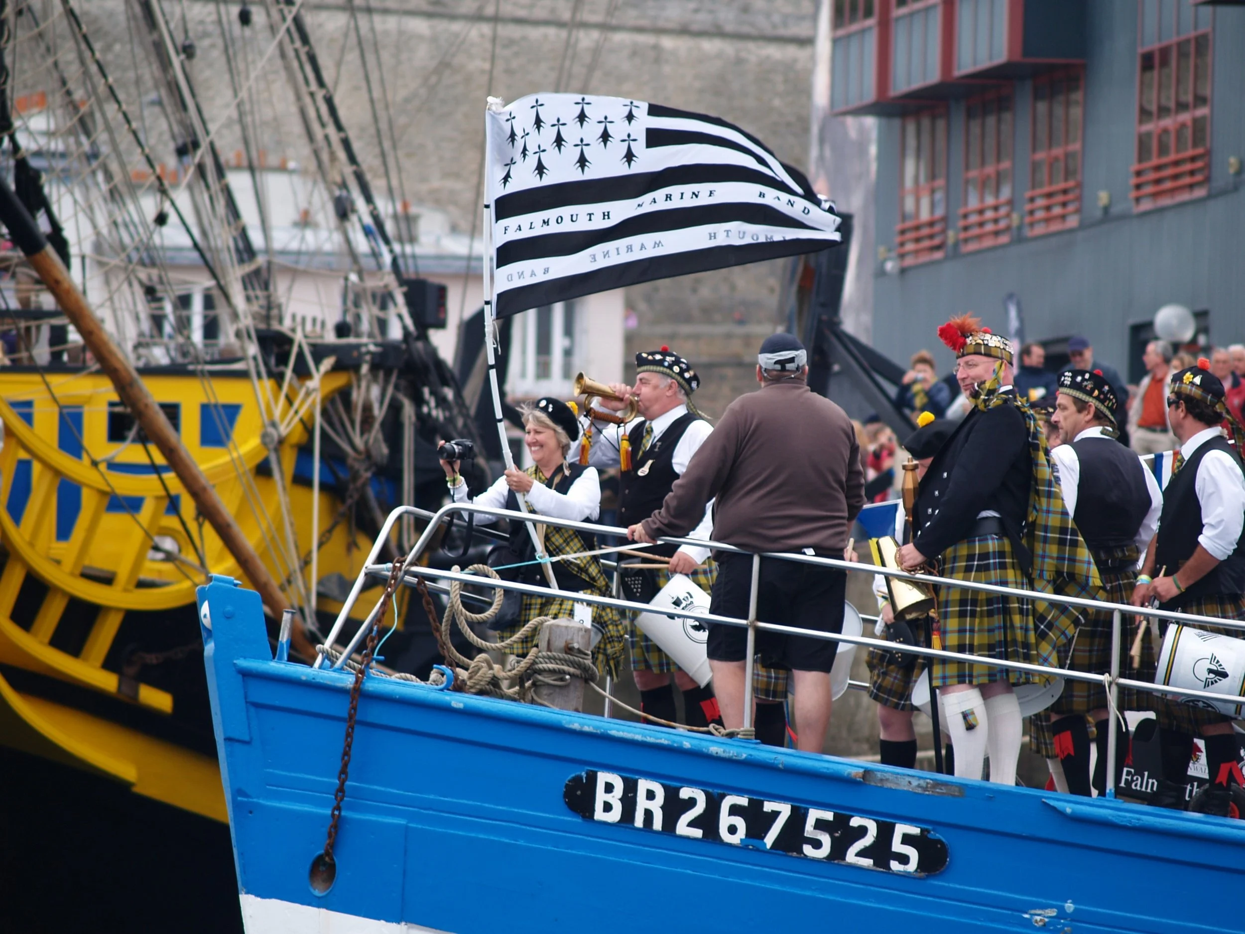 Groupe de personnes en vêtements traditionnels écossais, participant à un événement public avec une voile noire et blanche portant le nom 'Falmouth Marine Band'.