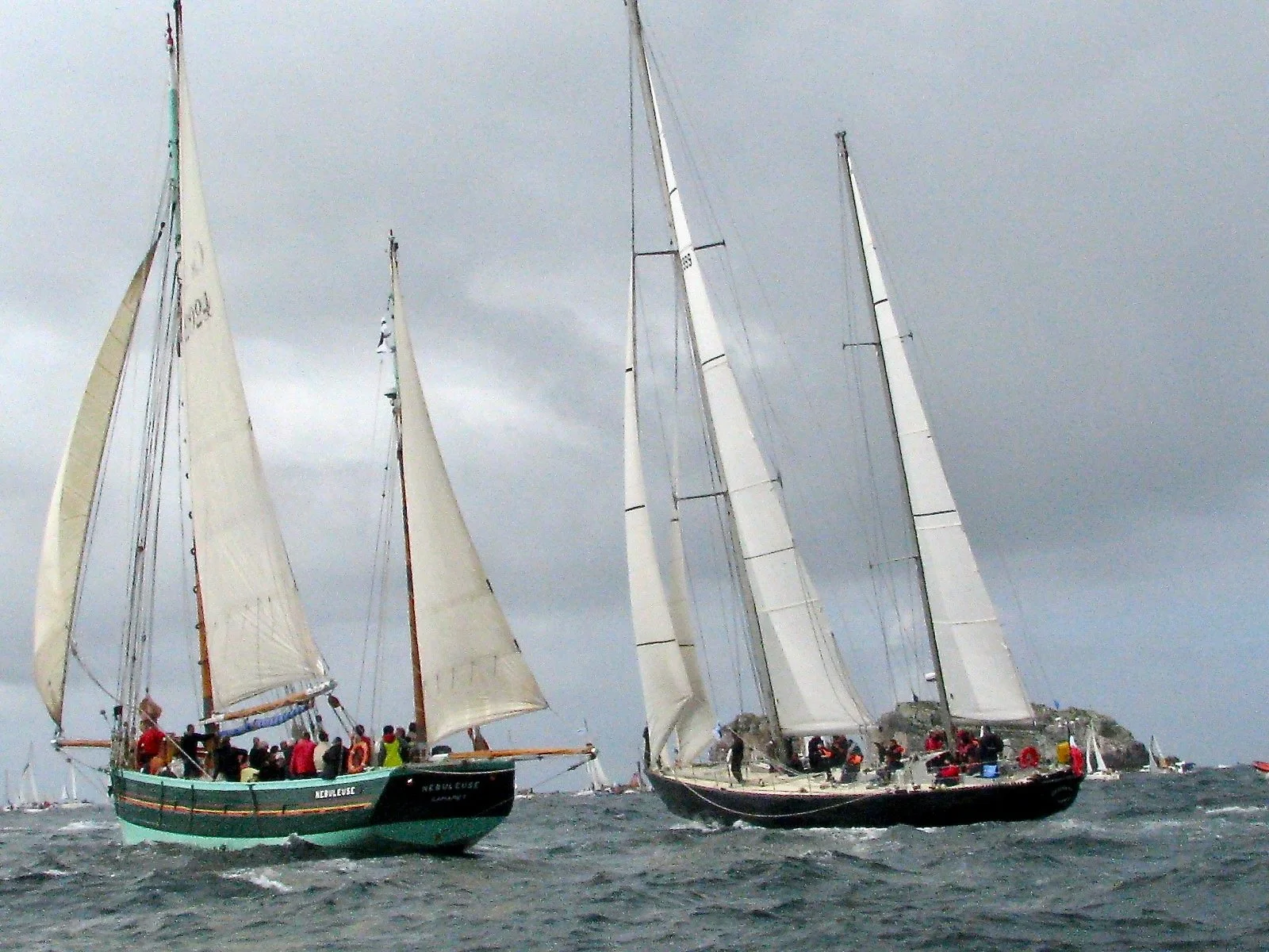 Deux voiliers en mer sous un ciel nuageux, avec des équipages à bord, naviguant près d'une côte rocheuse.