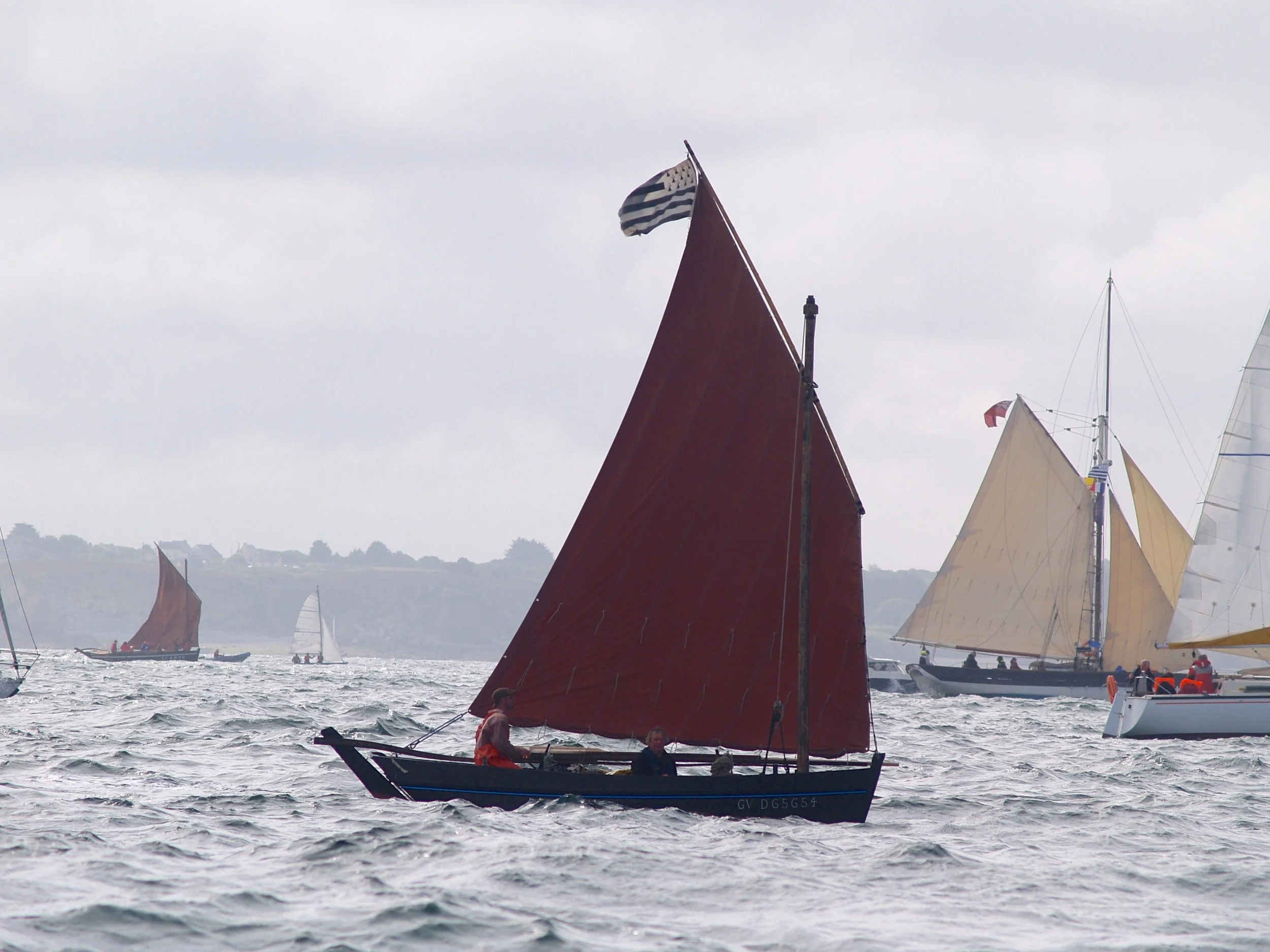 Voilier avec grande voile rouge et voisine avec voile beige, sur l'eau, avec plusieurs autres voiliers en arrière-plan, ciel nuageux, en mer.
