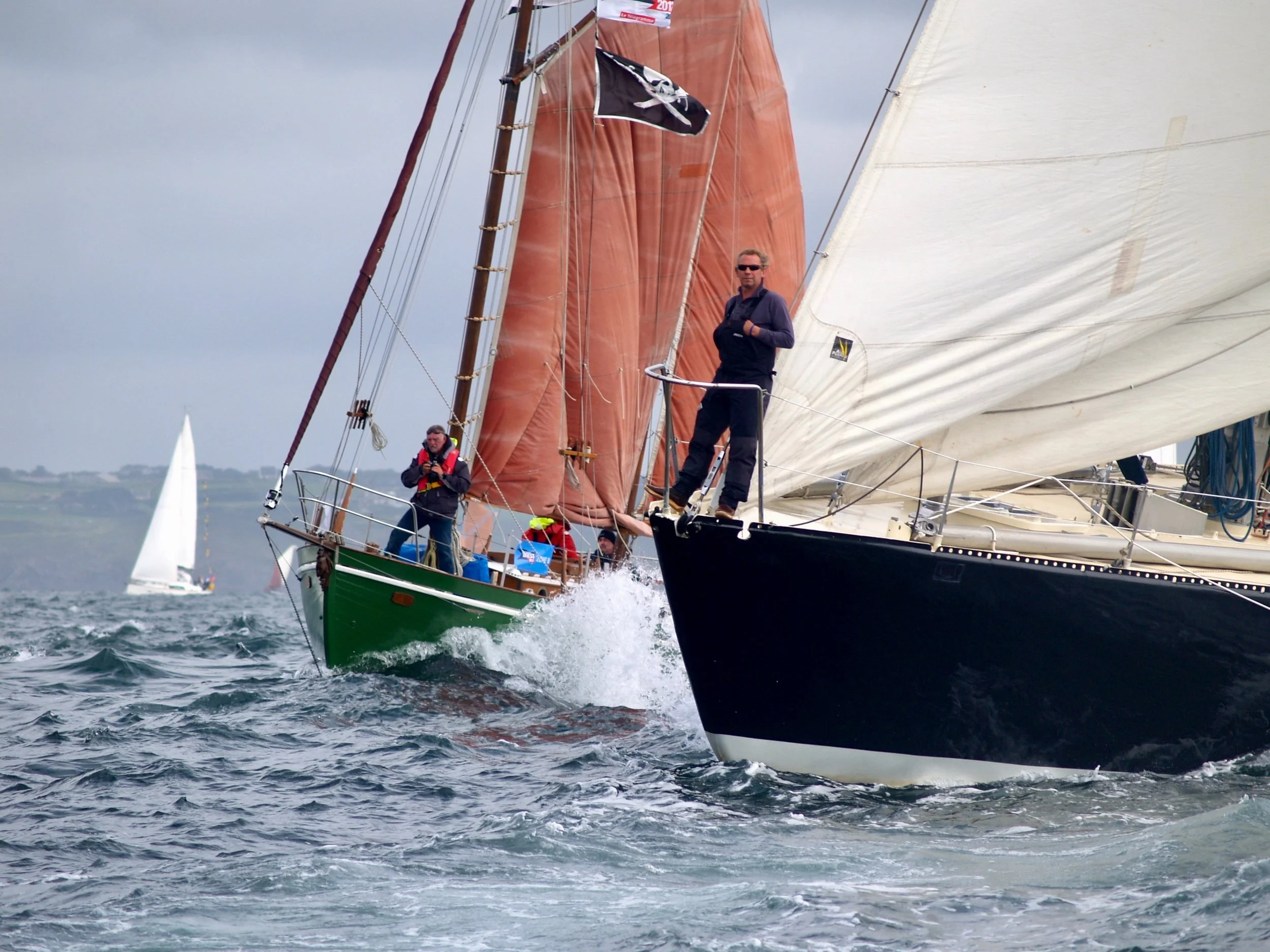 Une embarcation à voile naviguant sur l'eau agitée, avec deux personnes à bord, dont une debout en haut du bateau, portant des lunettes de soleil et un vêtement noir. Le voilier a des voiles colorées, une noire avec un crâne blanc, et une autre blanc
