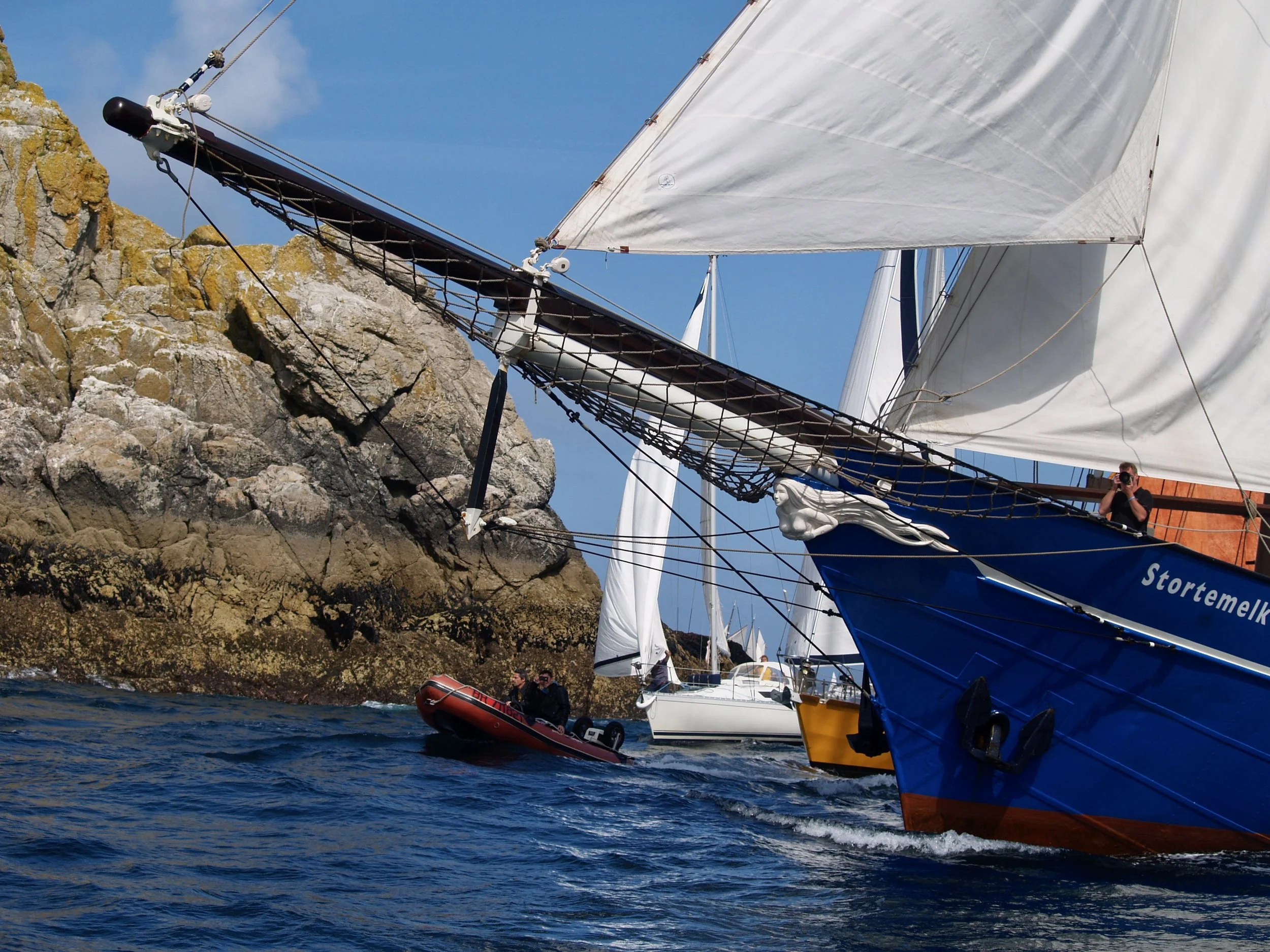 Un voilier blanc avec des voiles déployées navigue près de rochers, avec une personne dans une petite embarcation de secours à côté.