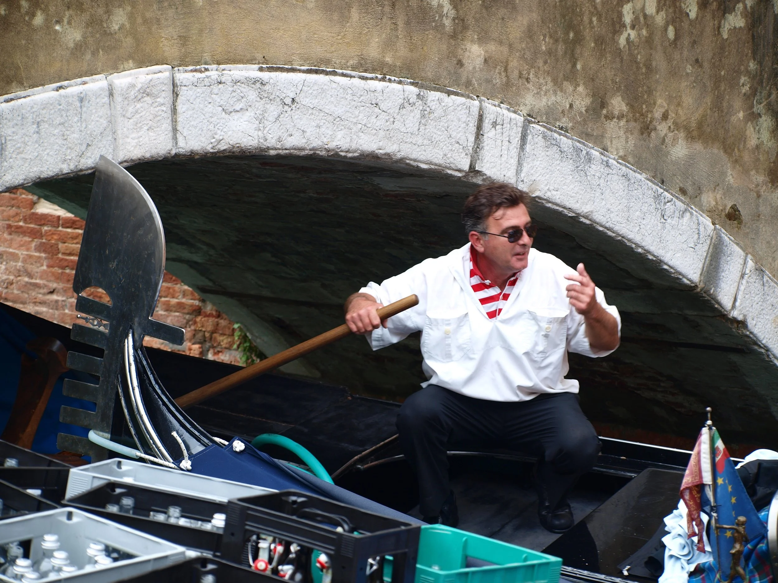 Un homme portant des lunettes noires, une chemise blanche et un t-shirt à rayures rouges et blanches, jouant avec un bâton près d'un bateau en plein air sous un pont en pierre.