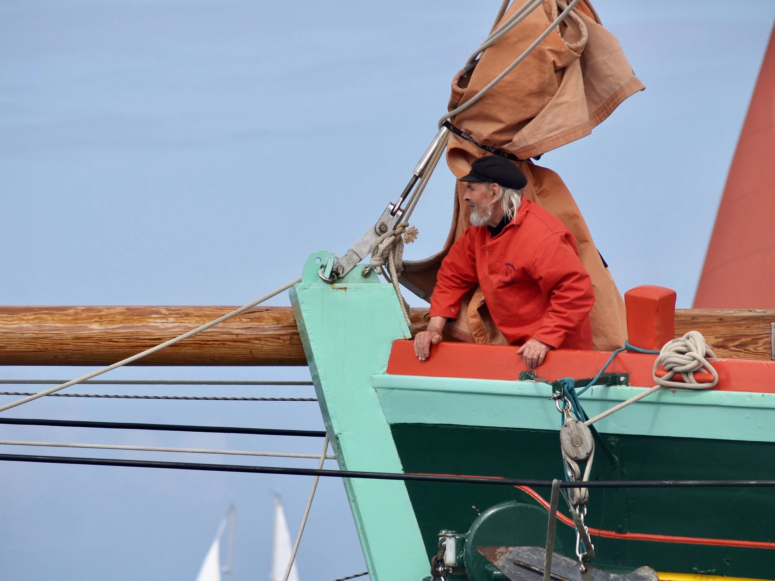 Un homme âgé portant une veste rouge et un bonnet noir est à bord d'un bateau, appuyé sur la rambarde. Le bateau est peint en vert clair et orange. Le fond montre un ciel bleu, avec un mat à voile blanc en arrière-plan.