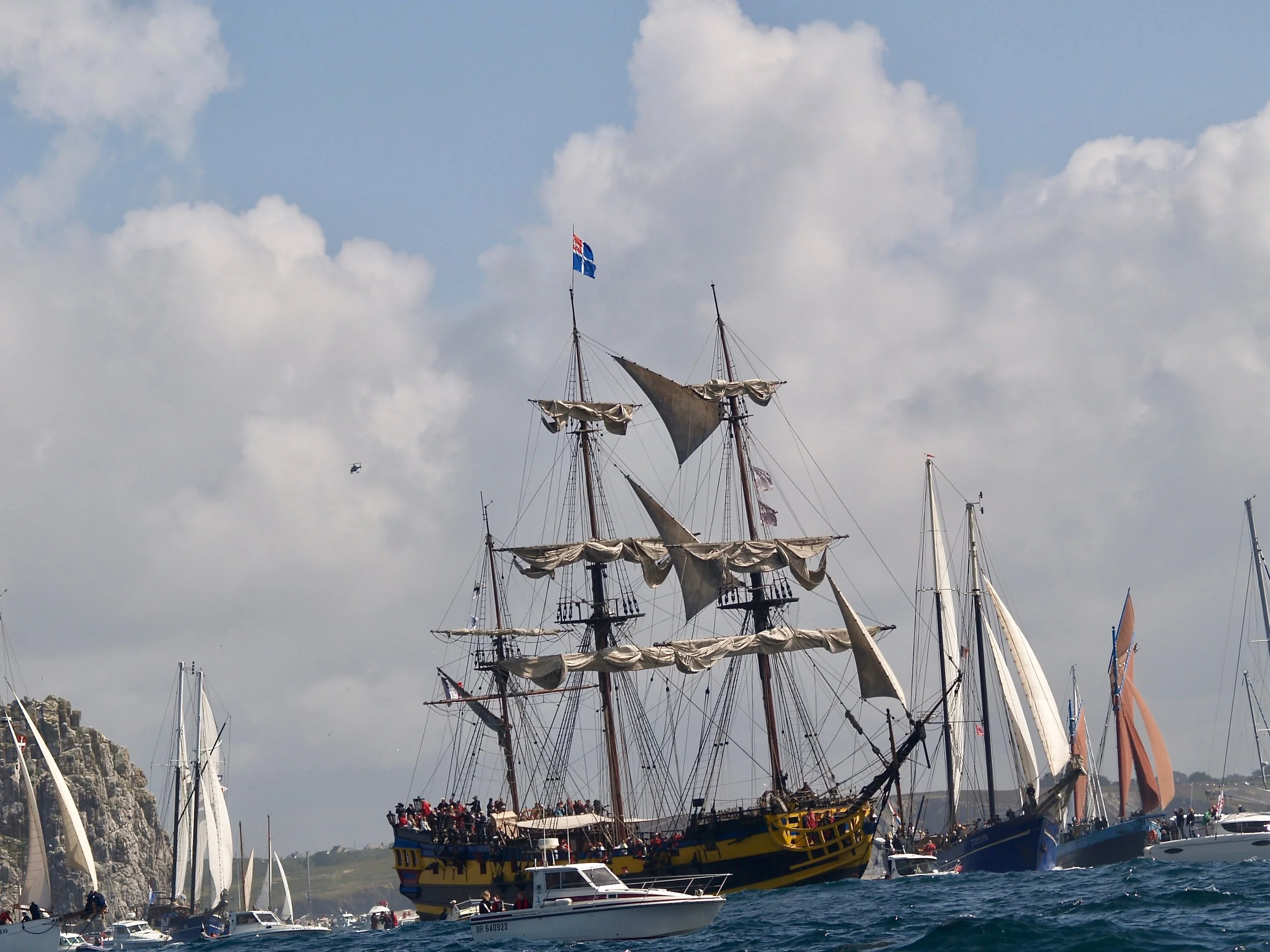 Un grand bateau de pirates avec des voiles en forme de pavillon noir et jaune navigue sur la mer, entouré de plusieurs petits yachts et voiliers.