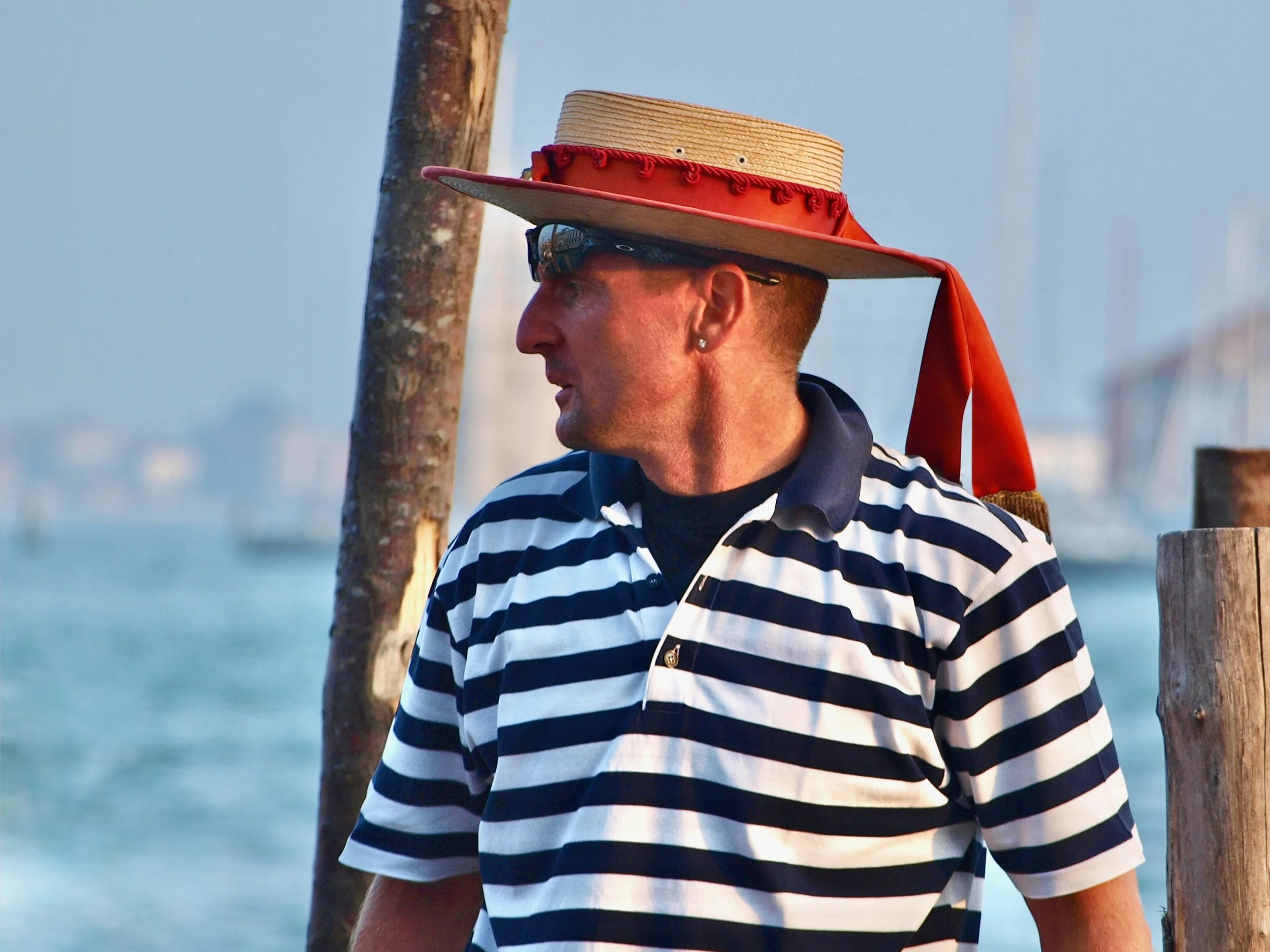 Un homme portant une chemise à rayures blanches et noires, un chapeau de style boater avec un ruban rouge, puis des lunettes de soleil, se tenant à côté d'un arbre en bois avec un plan d'eau en arrière-plan