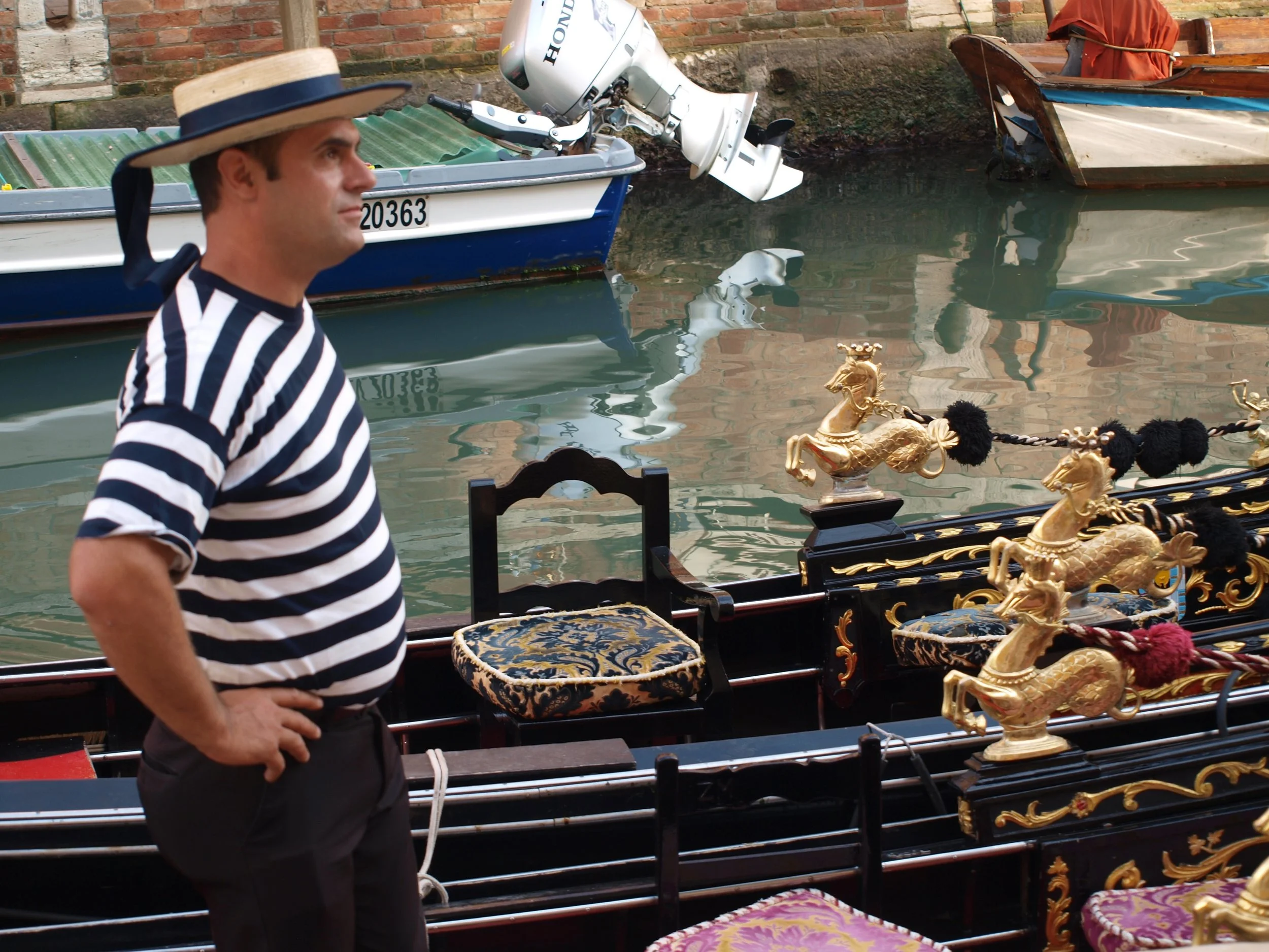 Un homme portant un chapeau et une chemise à rayures noires et blanches, debout à côté d'une gondole ornée de décorations dorées représentant des chevaux, dans un environnement aquatique avec des bateaux en arrière-plan.