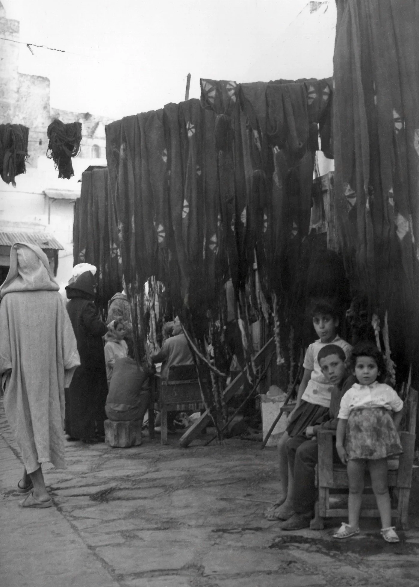 Des enfants et des adultes dans un marché en plein air, avec des vêtements suspendus comme décor ou étalage. Certains enfants sont assis sur des bancs, d'autres regardent ou discutent.