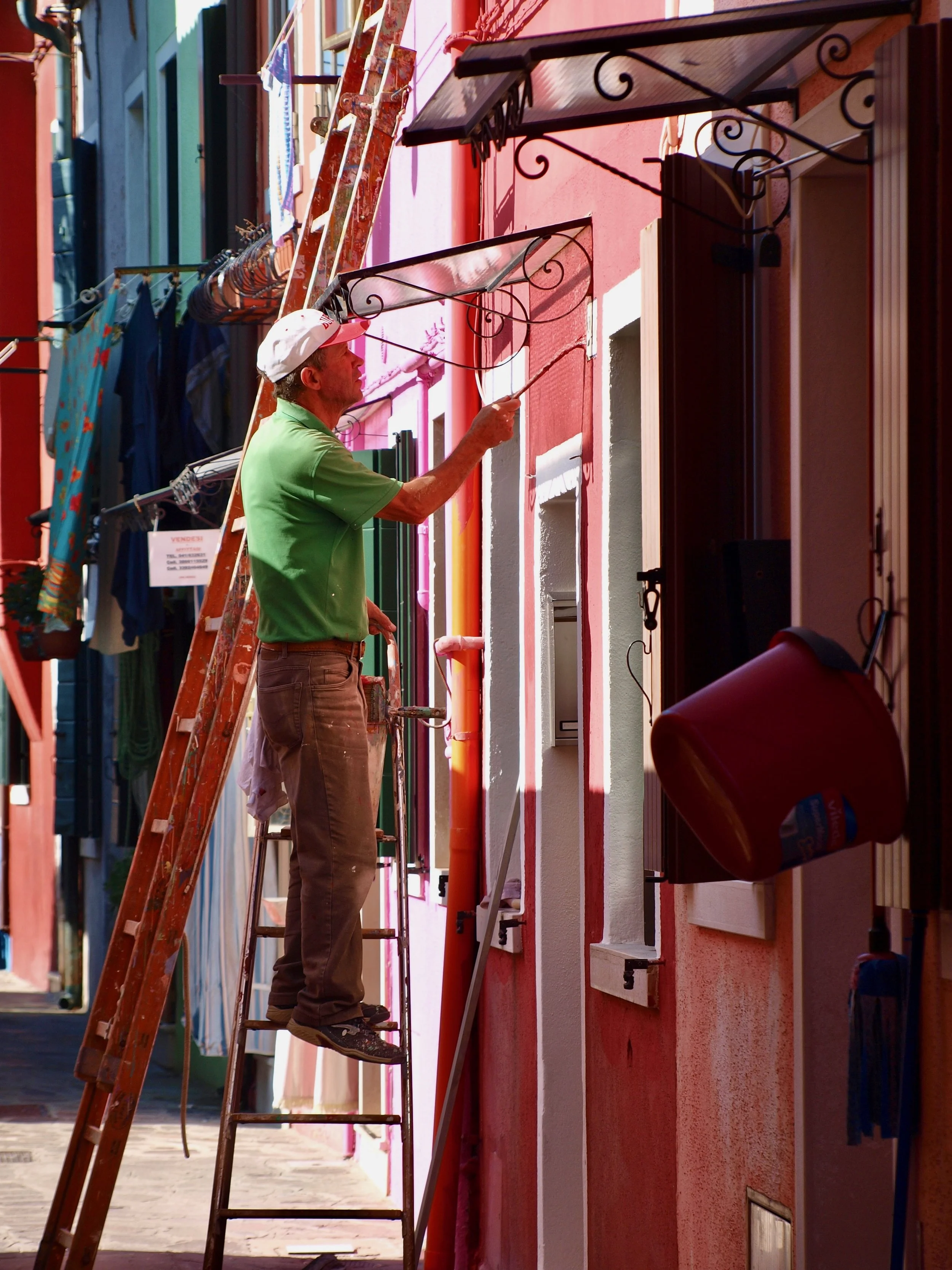 Un homme en polo vert et casquette blanche nettoie une façade de bâtiment colorée à l'aide d'un balai ou d'une brosse. Il se trouve sur une échelle en bois, et il y a une autre échelle en aluminium à côté de lui. La façade du bâtiment est rose avec d