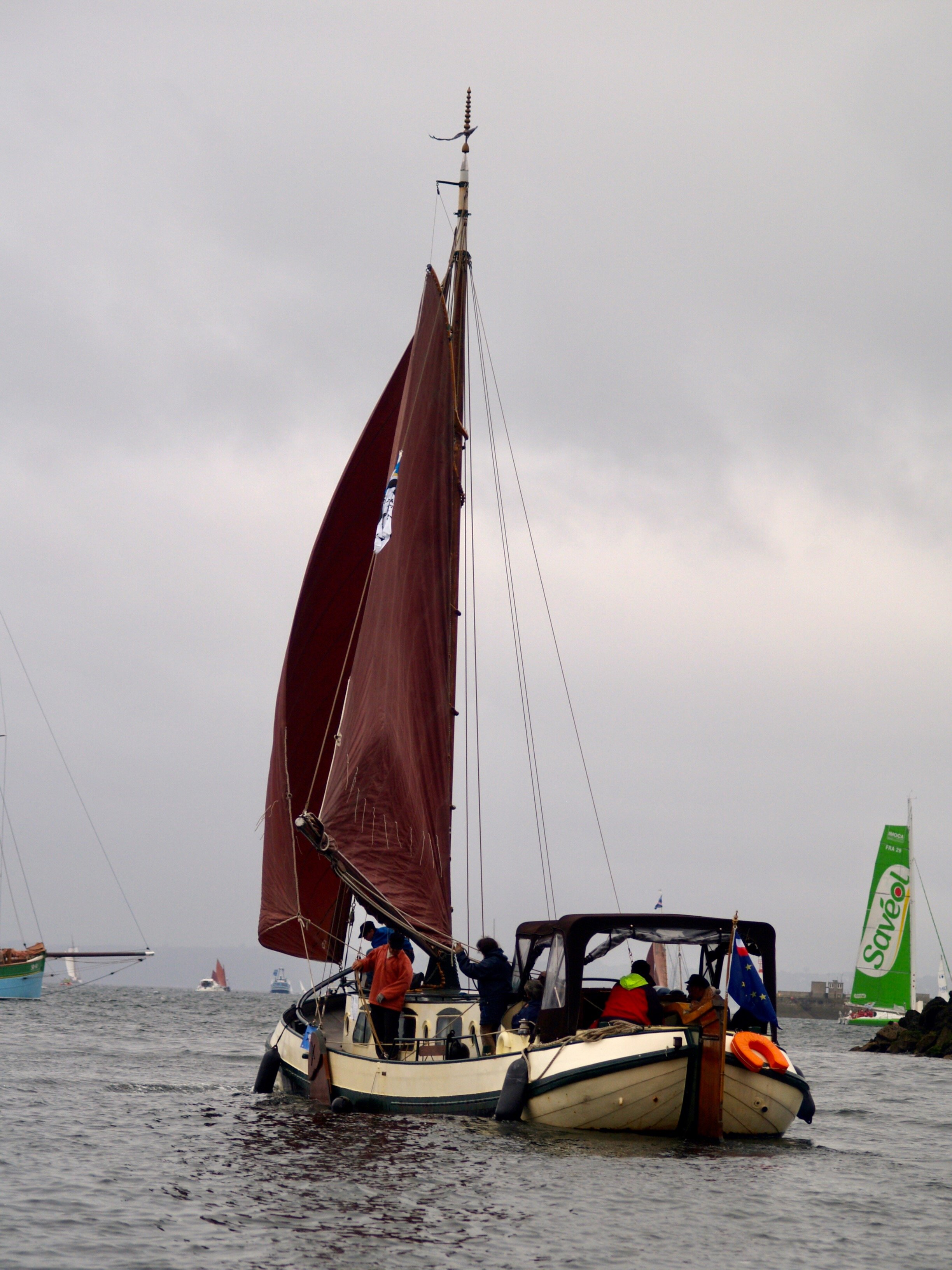 Un voilier naviguant sur l'eau, avec plusieurs personnes à bord, des autres bateaux et voiliers en arrière-plan, et des drapeaux multicolores, dans un ciel nuageux.