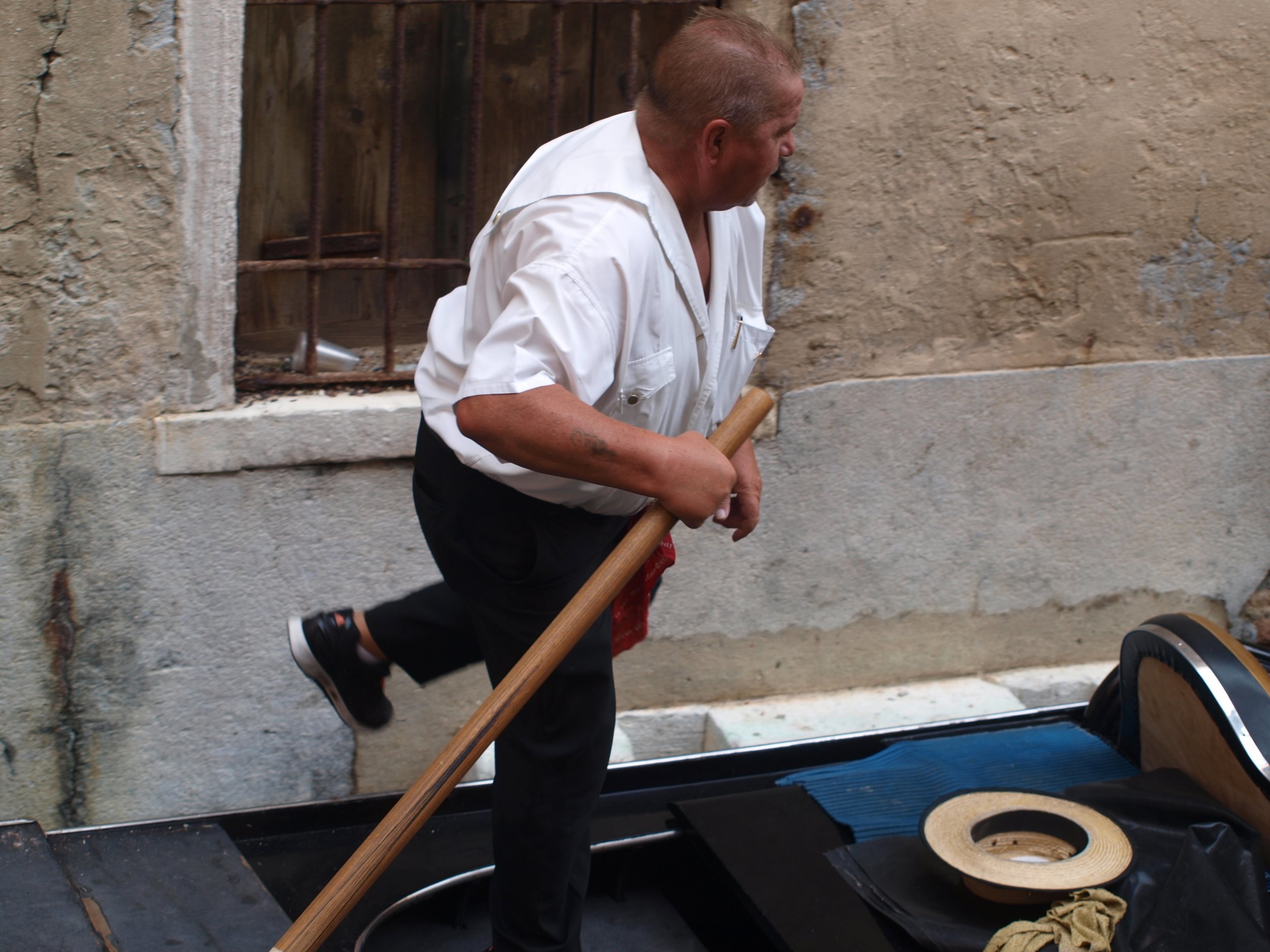 Un homme âgé, vêtu d'une chemise blanche à manches longues, marche dans une rue pavée avec un bâton à la main, à côté d'une vieille maison en pierre avec une fenêtre grillagée en fer.