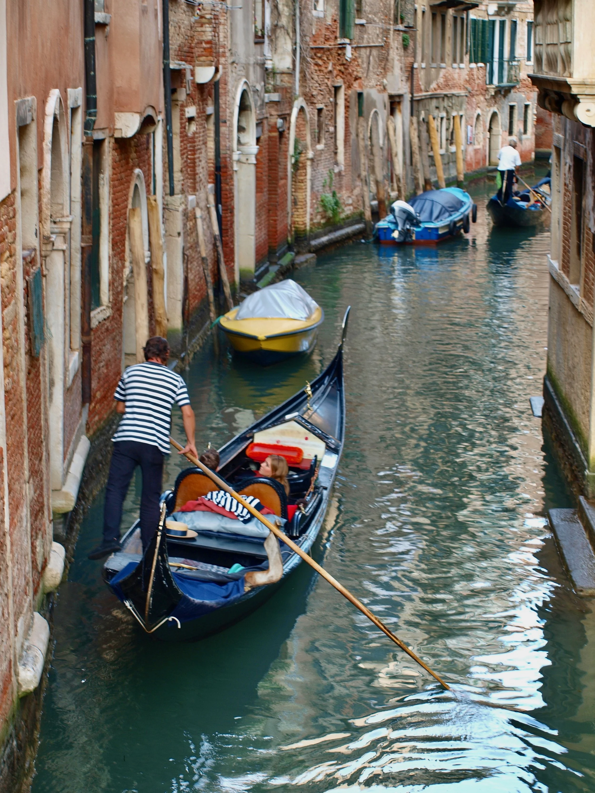 Une gondole voguant dans un canal étroit bordé de bâtiments en briques et en pierre à Venise, avec un gondolier et des touristes à bord.