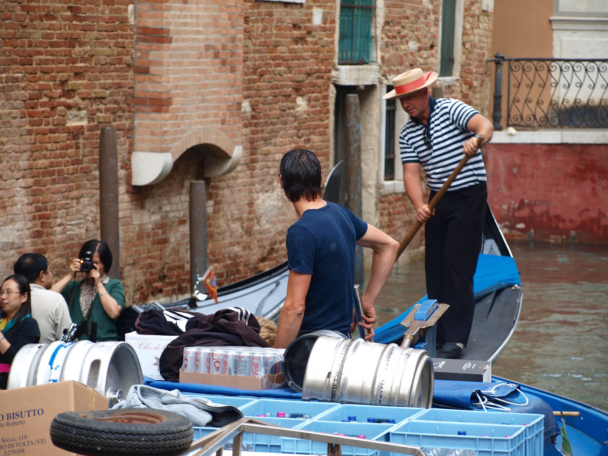 Un homme portant un chapeau de paille à rayures bleues et blanches, en train de parler avec un jeune homme avec des lunettes, dans une rue pavée avec un mur en briques en arrière-plan. Divers objets recyclés et boîtes sont visibles en premier plan.