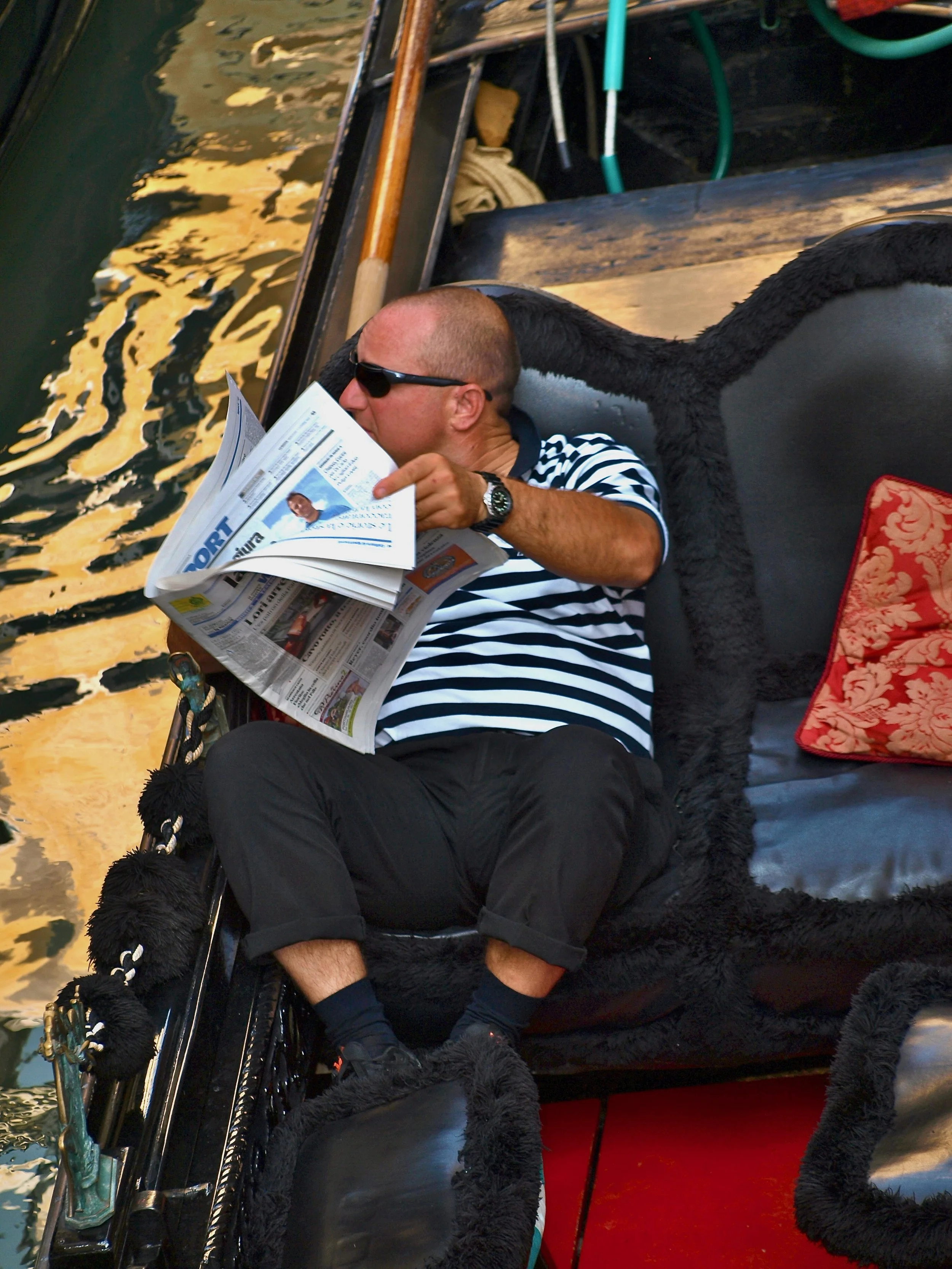 Homme en tenue rayée de noir et blanc, portant des sunglasses, assis sur une gondole, lisant un journal, avec un décor de gondole vénitienne en arrière-plan.
