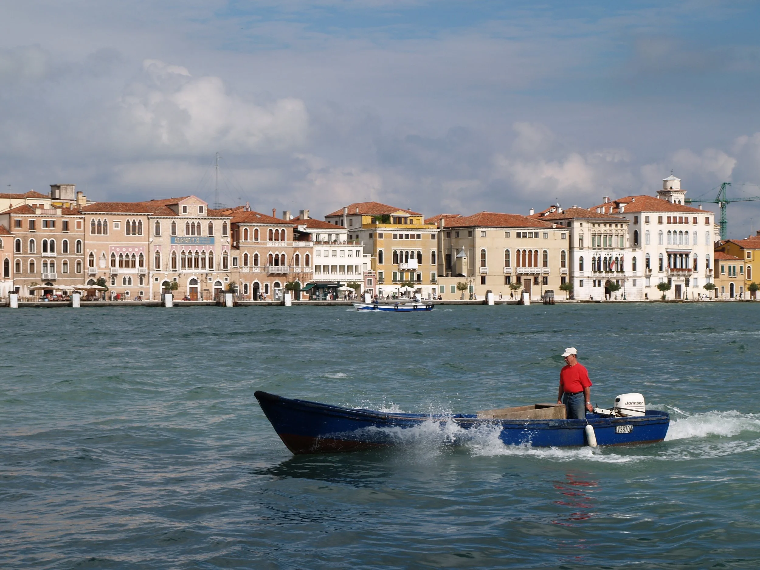 Une scène de ville italienne avec des bâtiments colorés au bord de l'eau, un bateau à moteur noir en premier plan, un homme en t-shirt rouge et chapeau blanc sur le quai.