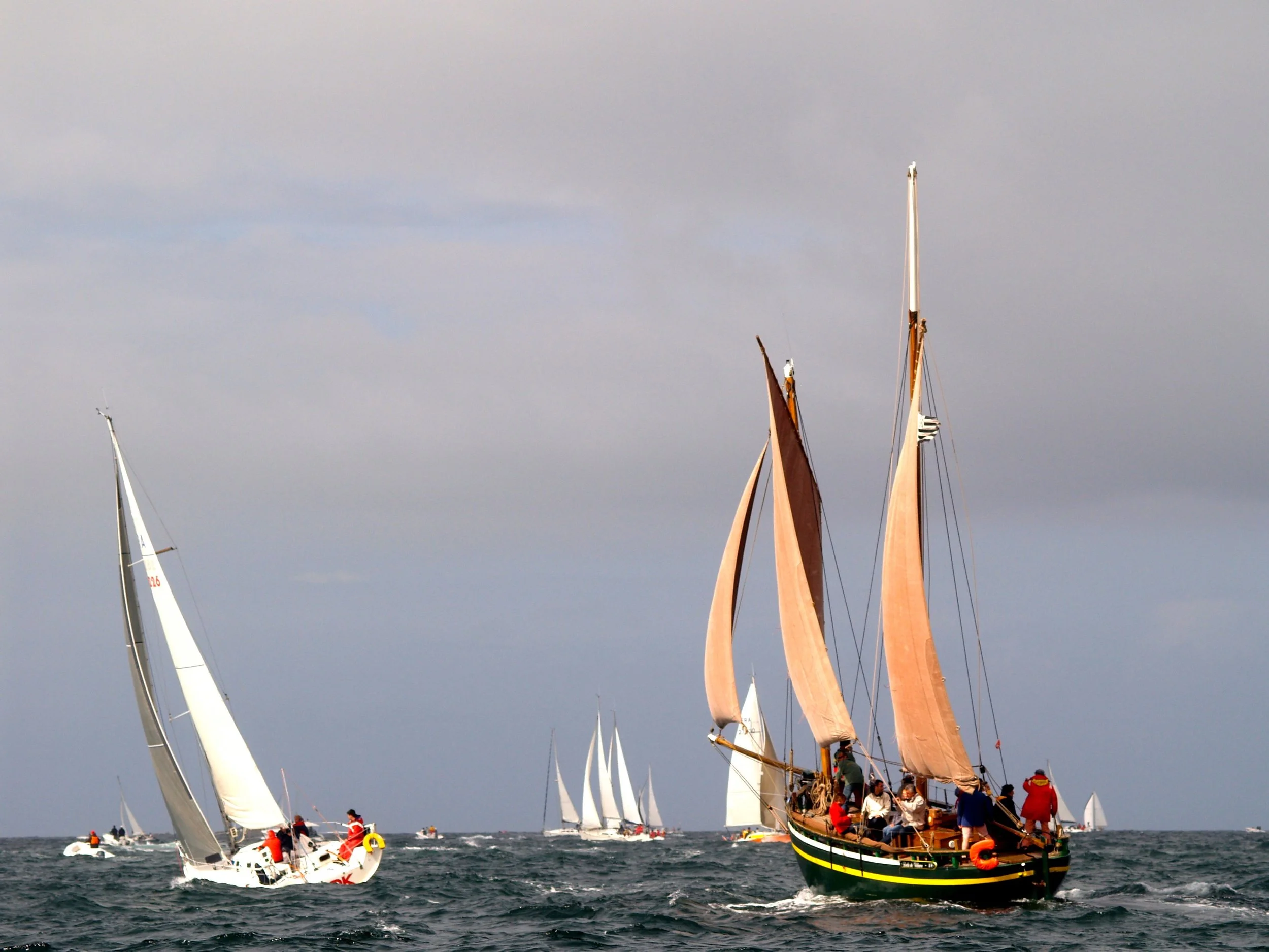 Plusieurs voiliers avec des voiles blanches et colorées naviguent sur la mer sous un ciel nuageux.