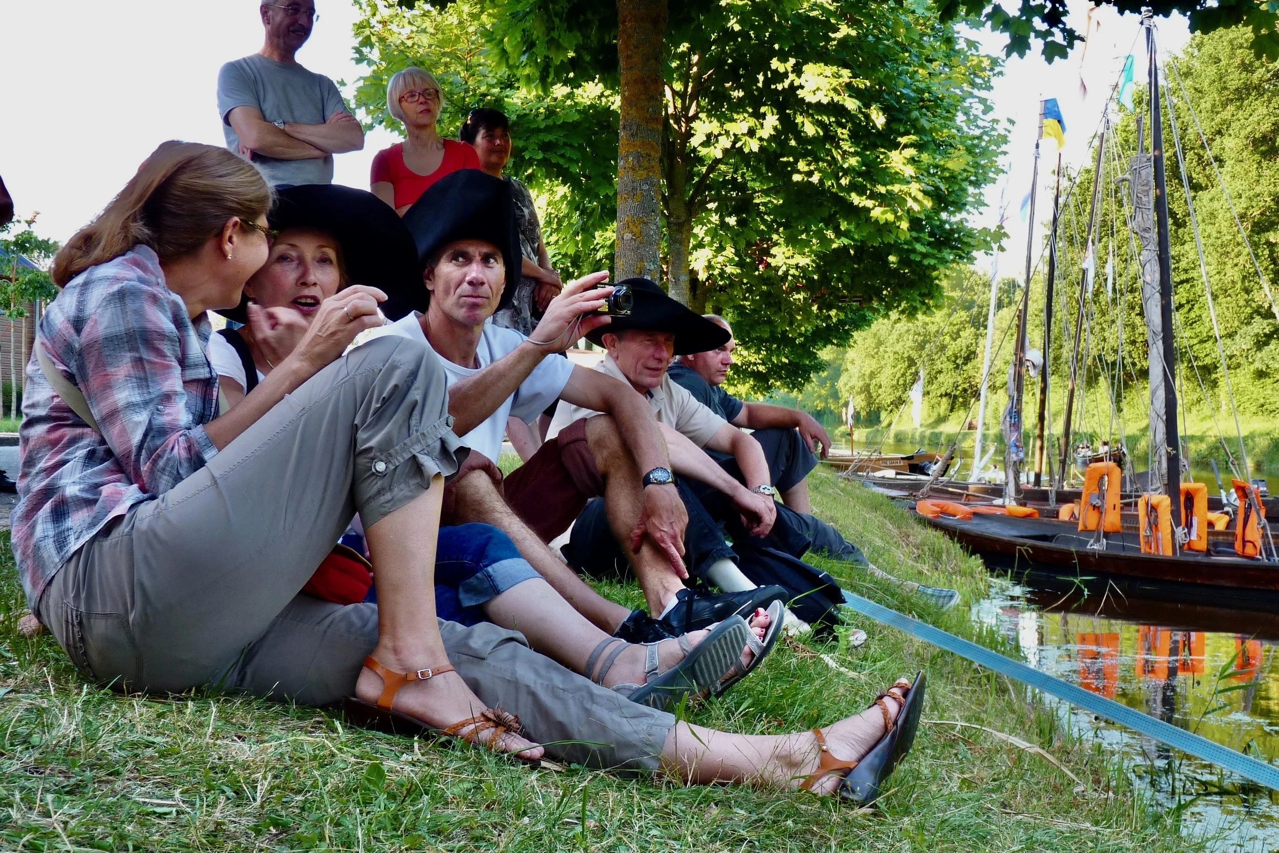 Un groupe de personnes assises et debout près d’un lac avec des bateaux. Certaines portent des chapeaux, d’autres des vêtements décontractés, et ils semblent discuter ou observer quelque chose sur l’eau. Il y a des arbres verts en arrière-plan.
