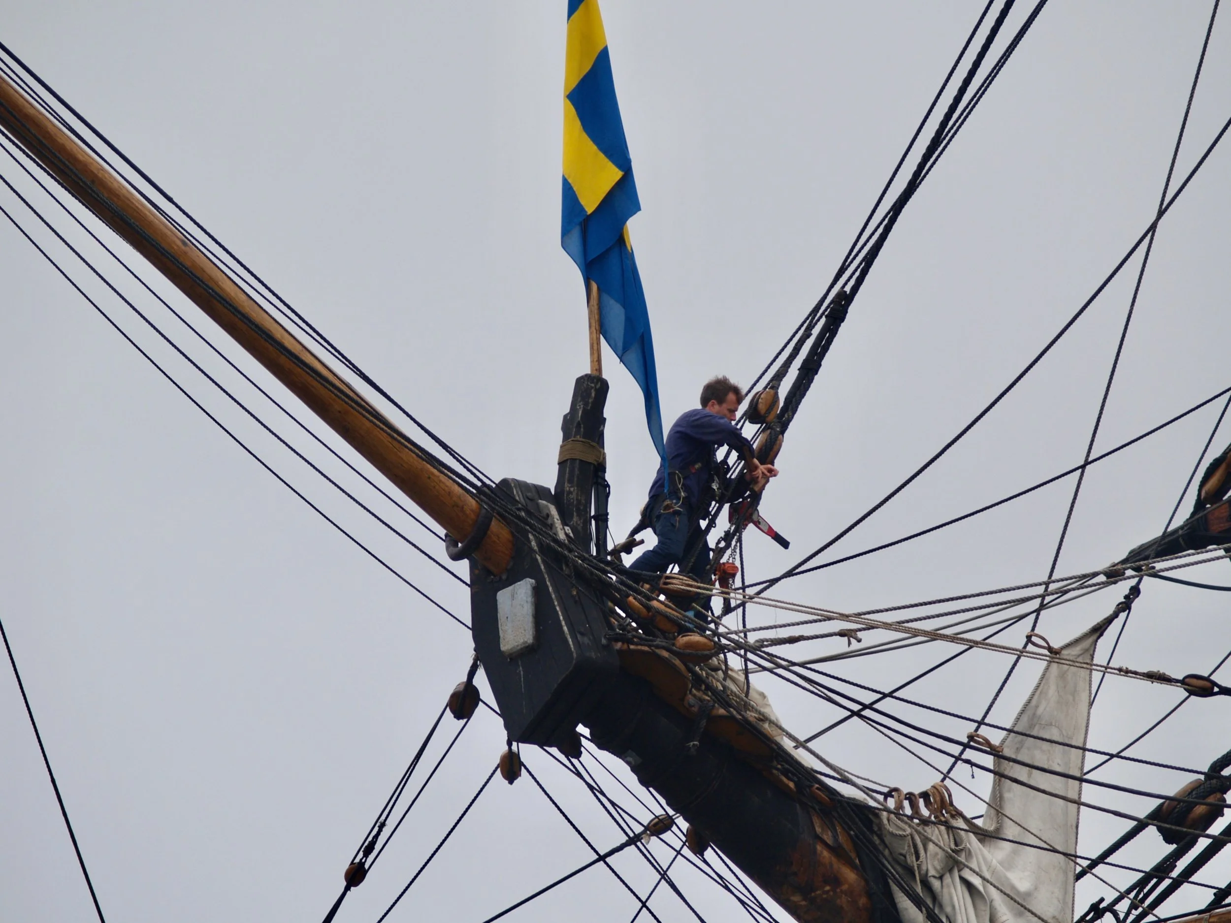 Un homme travaillant à la proue d'un voilier, attaché à une mât et au gréement, avec un drapeau bleu et jaune flottant au sommet, vu de dessous contre un ciel gris.