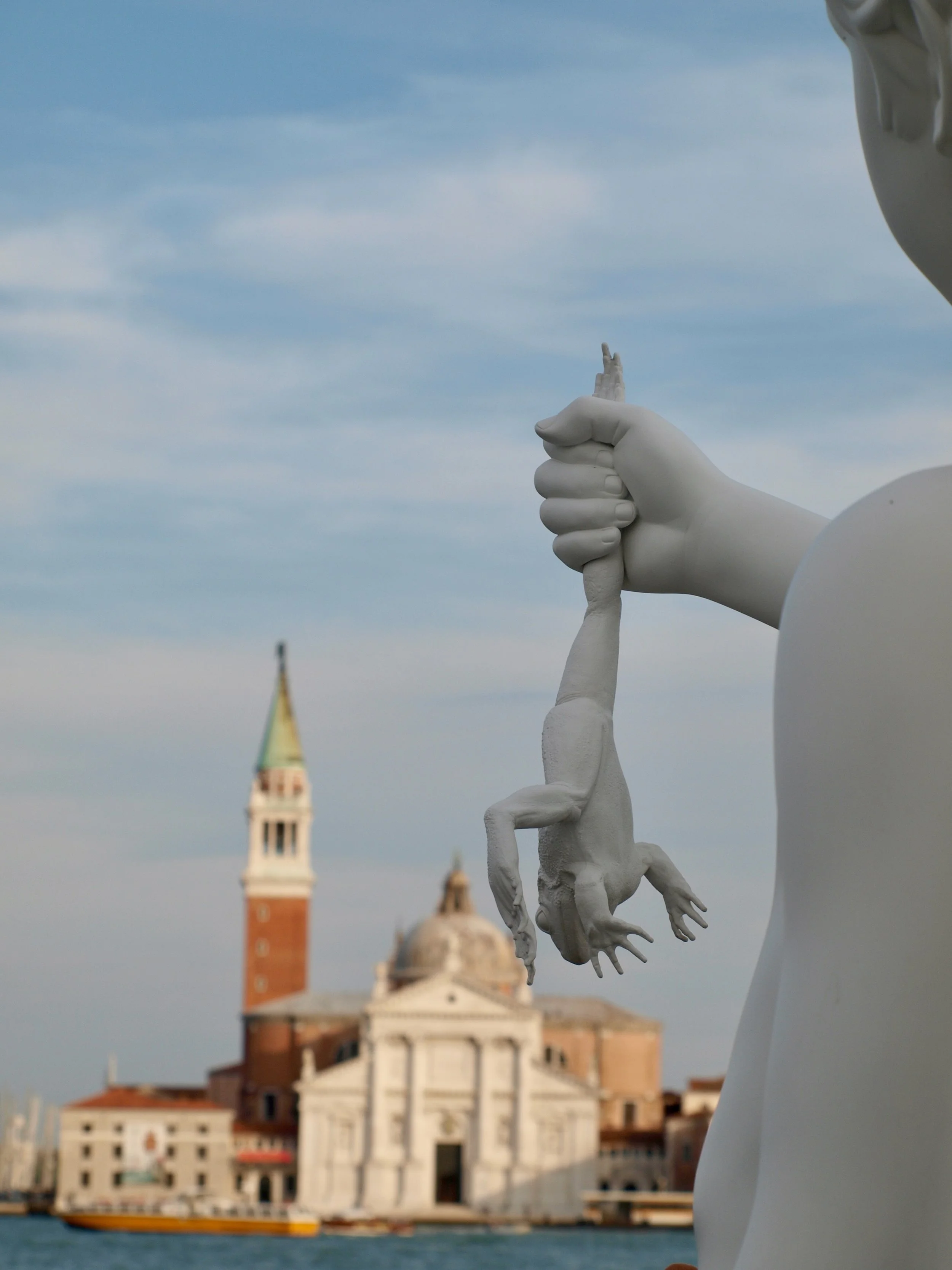 Photo d'une sculpture blanche représentant une main tendue, avec une vue de Venise en arrière-plan, incluant le Campanile de San Marco et le canal avec un bateau.