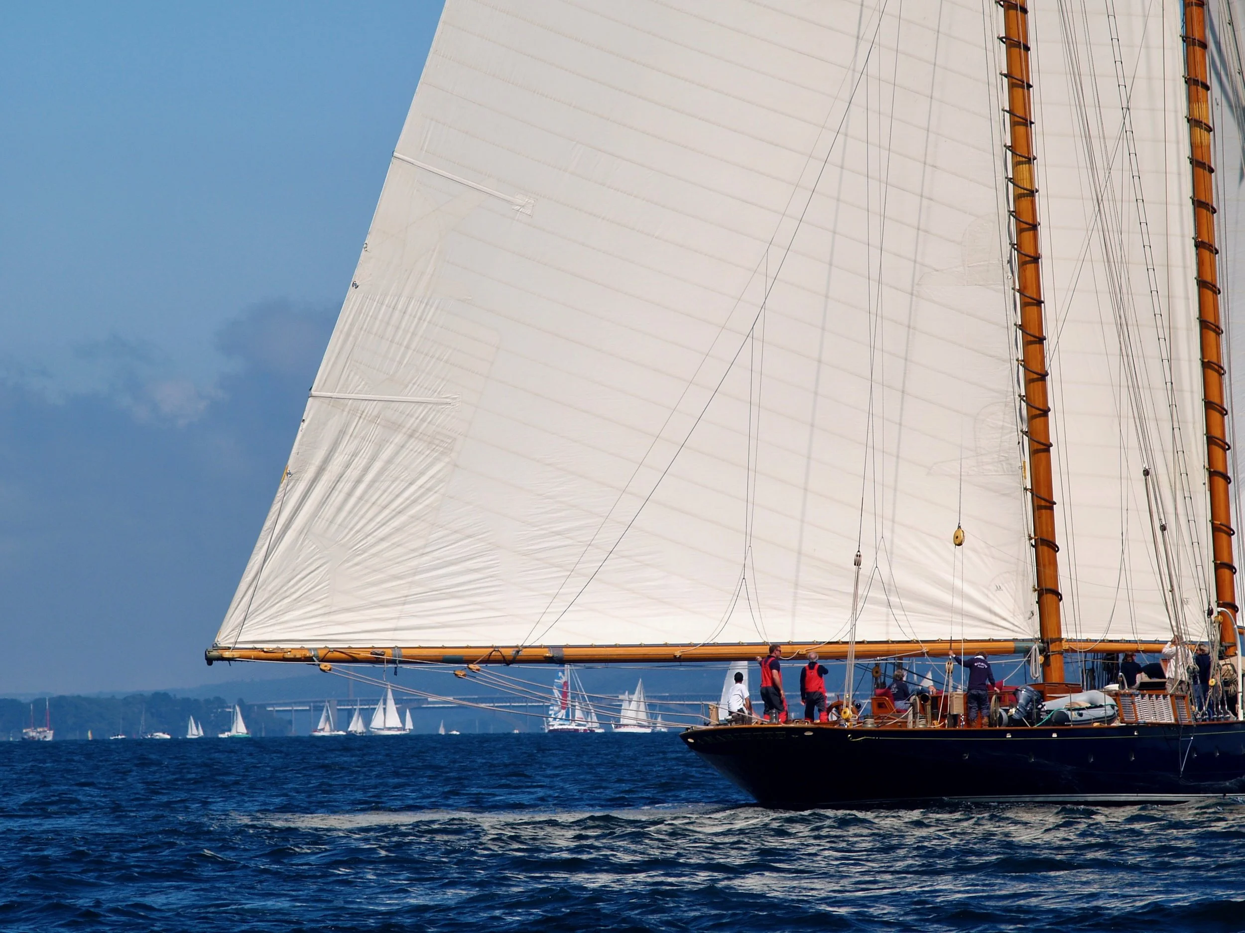 Un voilier à voile blanche avec plusieurs personnes à bord, naviguant sur la mer lors d'une journée ensoleillée avec ciel bleu.
