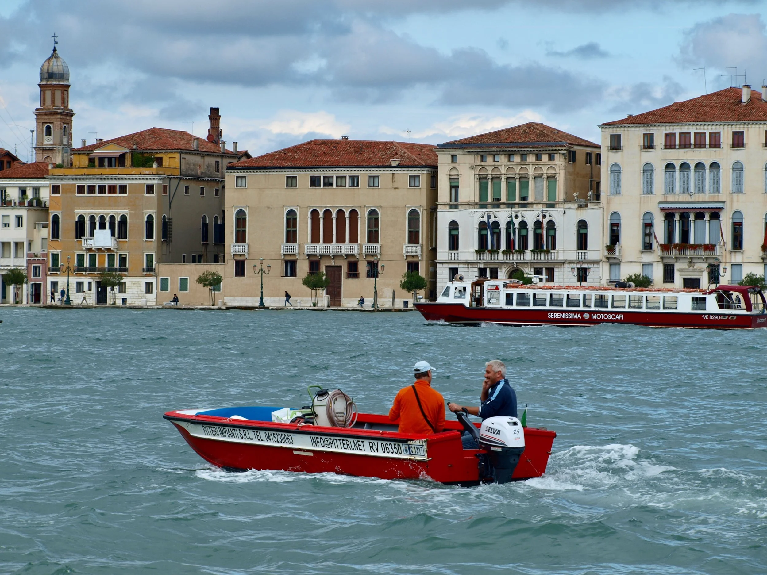 Deux hommes dans un bateau rouge naviguant sur l'eau, avec un ensemble de bâtiments anciens en arrière-plan et un ciel nuageux.