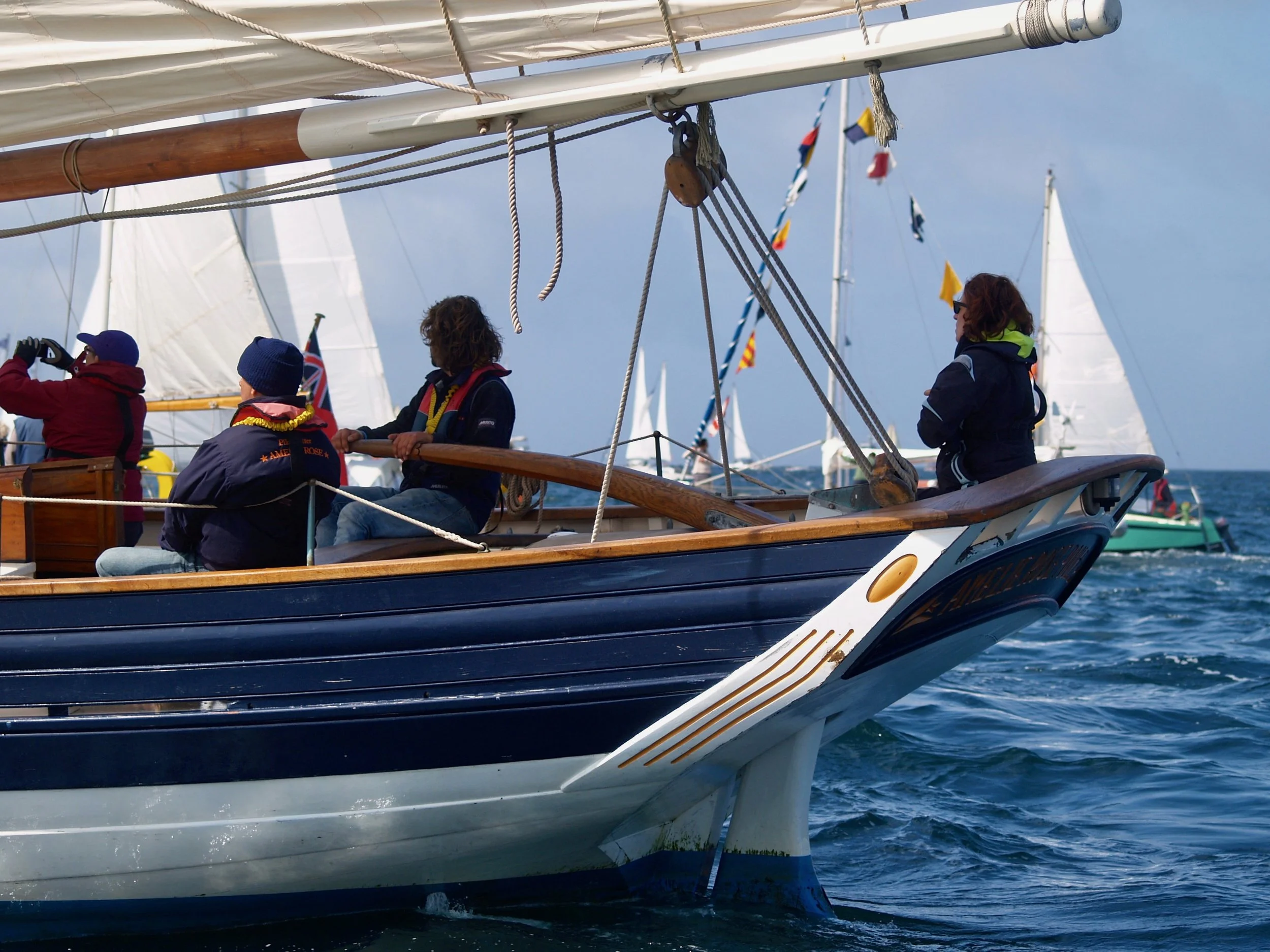 Trois personnes sur un voilier naviguant sur la mer, avec des voiles blanches et des cordages visibles, portant des vêtements chauds et un ciel en partie nuageux en arrière-plan.