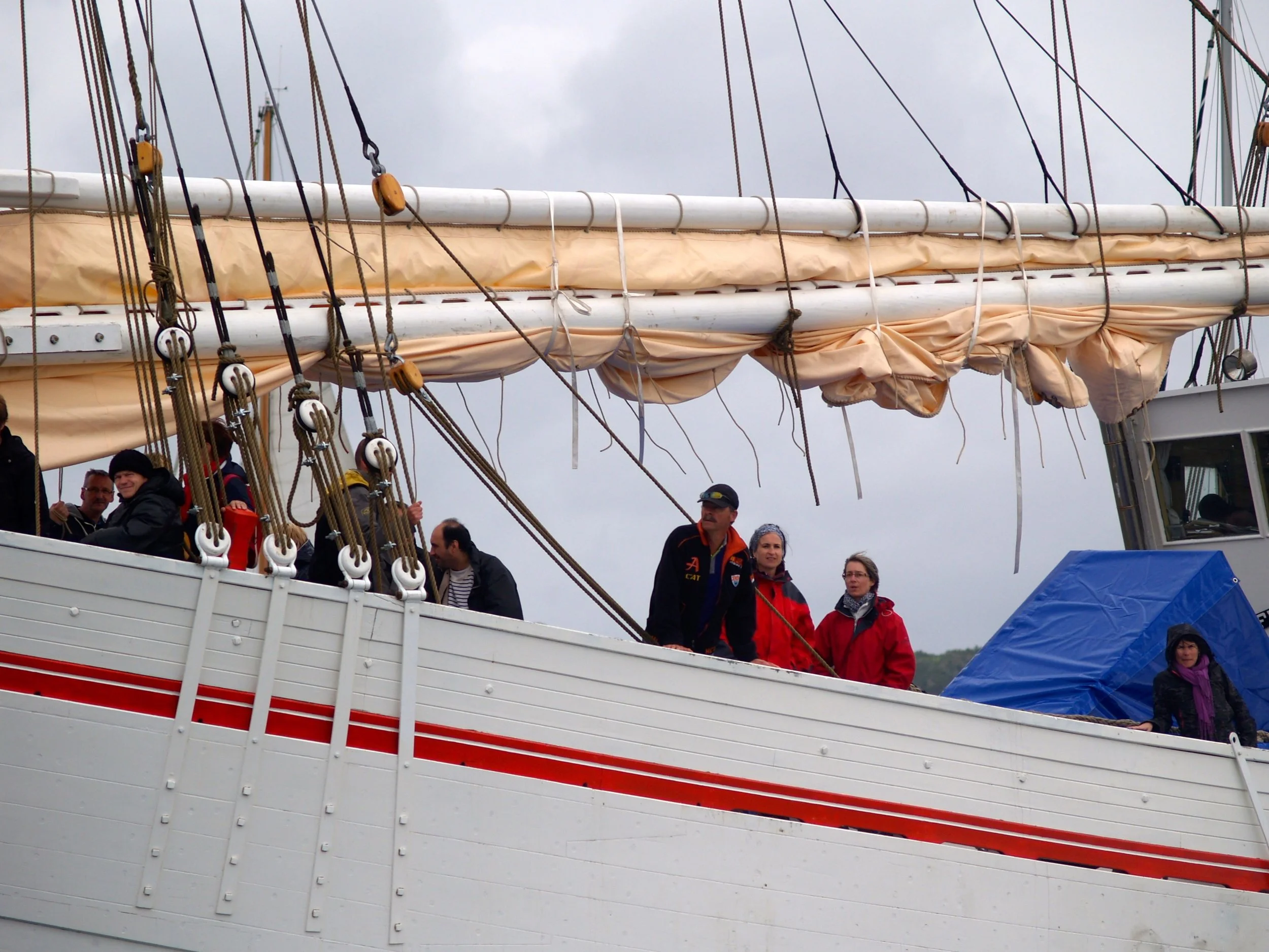 Groupe de personnes à bord d'un voilier avec plusieurs cordages, voiles repliées, et un ciel nuageux en arrière-plan.