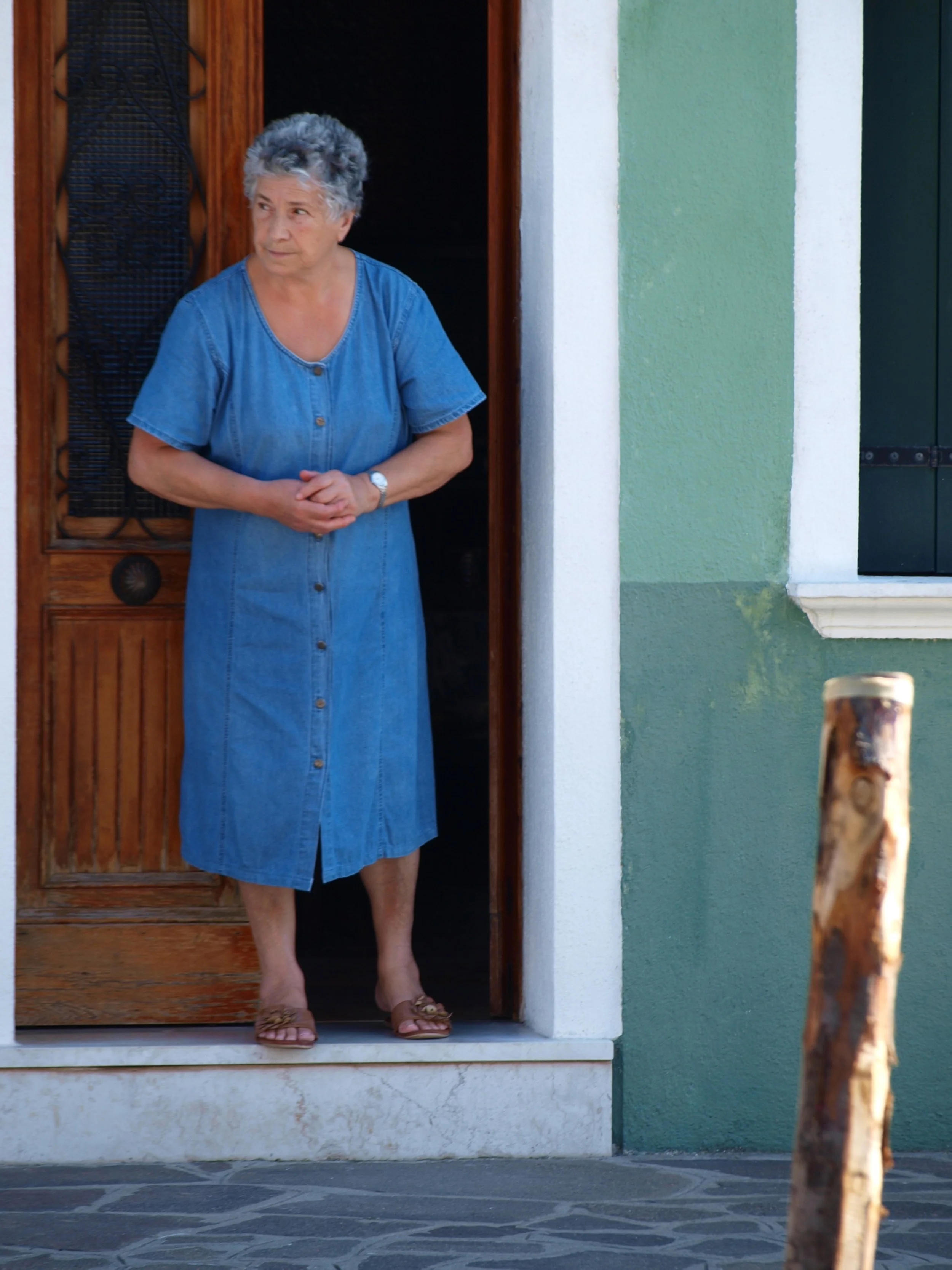 Une femme âgée, aux cheveux gris, porte une robe en jean bleu, se tient dans le cadre d'une porte en bois, devant un mur vert avec une fenêtre noire.