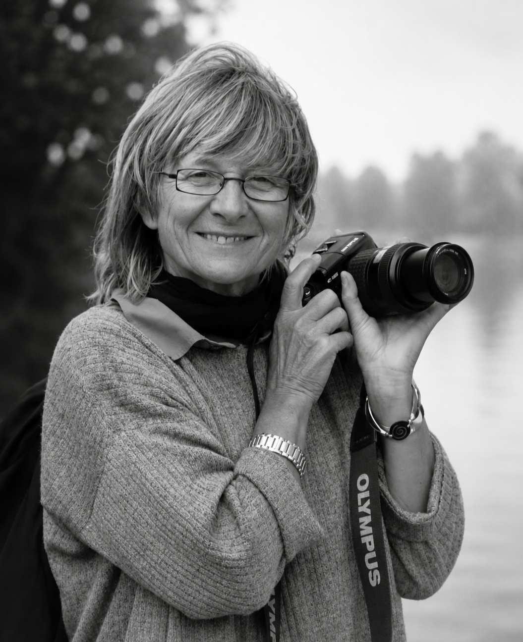 Femme âgée tenant un appareil photo numérique, souriant, portant des lunettes, un pull gris et une montre en argent, à l'extérieur près de l'eau.