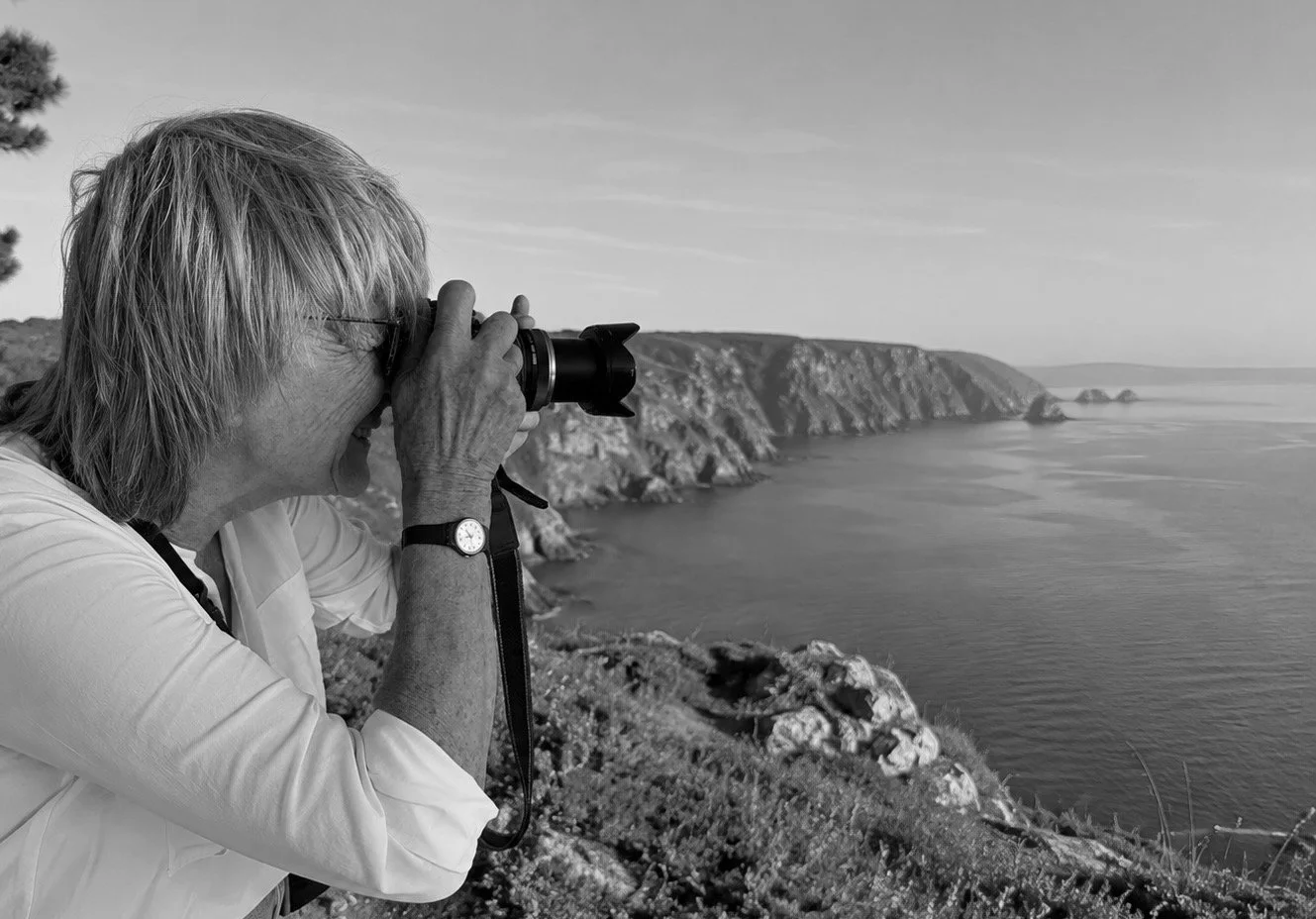 Une femme âgée avec des cheveux gris, portant une montre, prend une photo avec un appareil photo numérique en direction de la mer et des falaises sur une côte rocheuse. La scène est en noir et blanc.