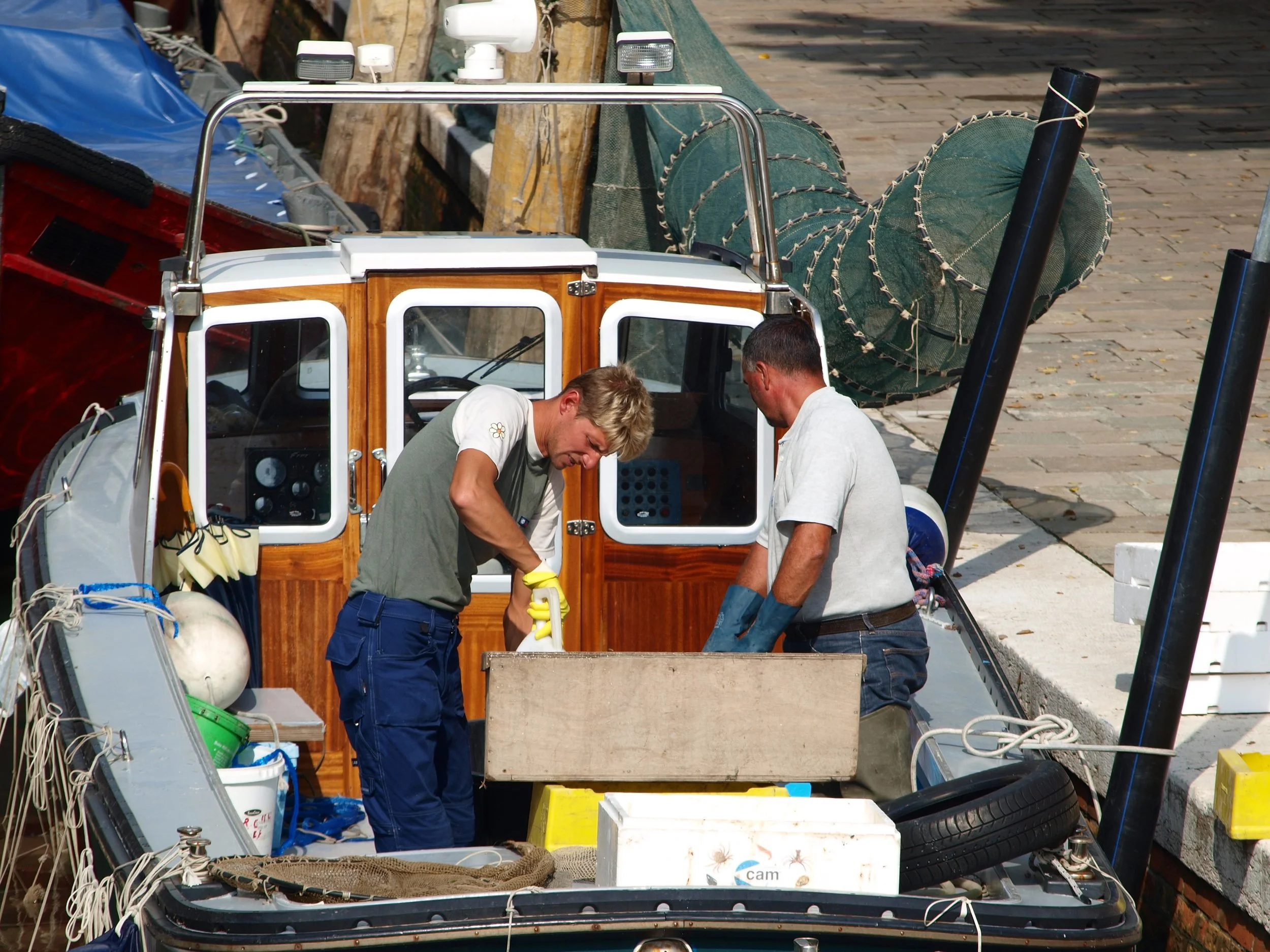 Deux hommes travaillent sur un bateau, qui comporte des éléments en bois et en métal, avec des équipements divers comme des roues, un câblage et une pompe. Le bateau est stationné sur un quai pavé.