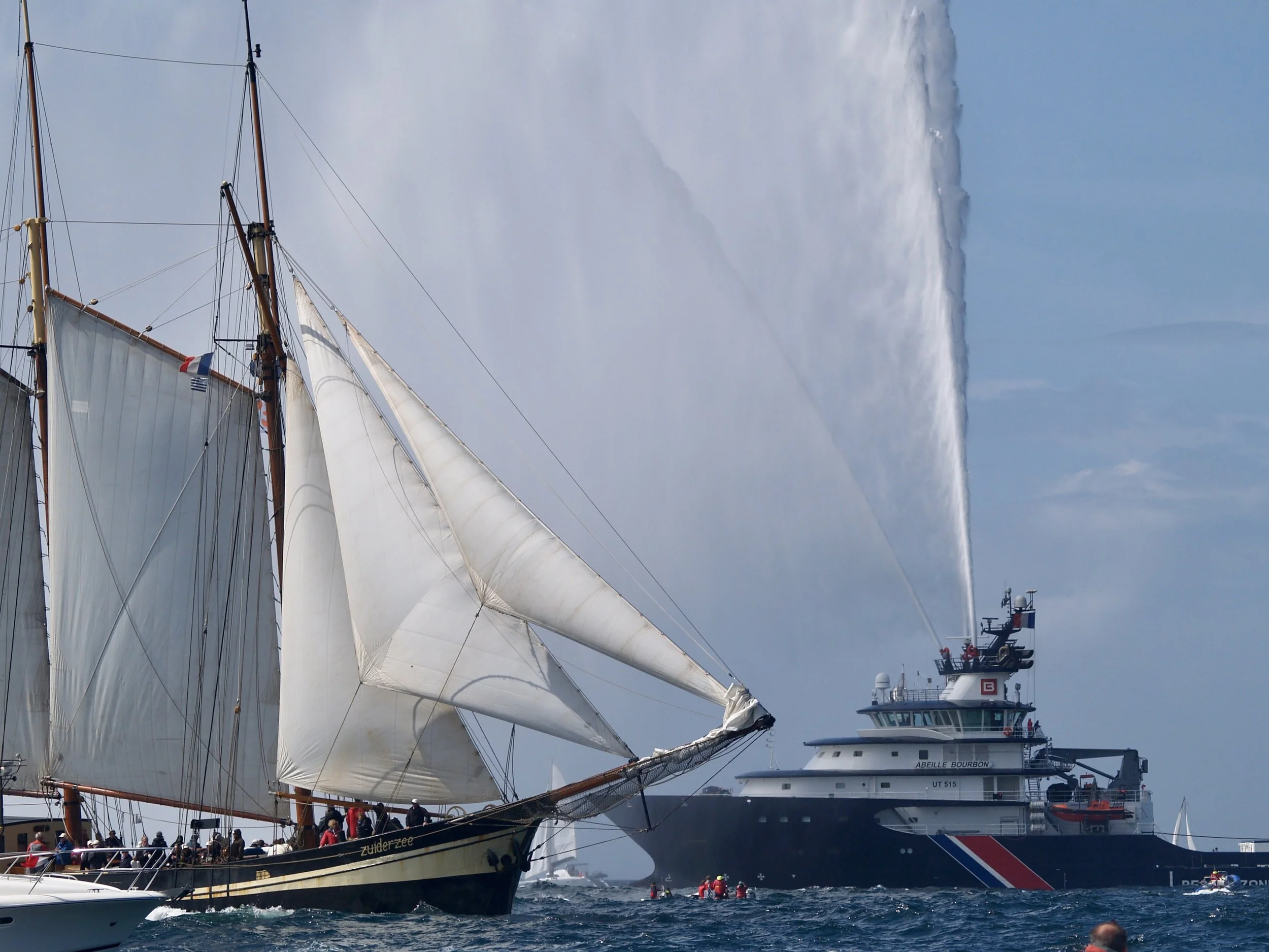 Un voilier avec des voiles déployées dans un port, un grand bateau de croisière en arrière-plan, en mer, avec un ciel nuageux.