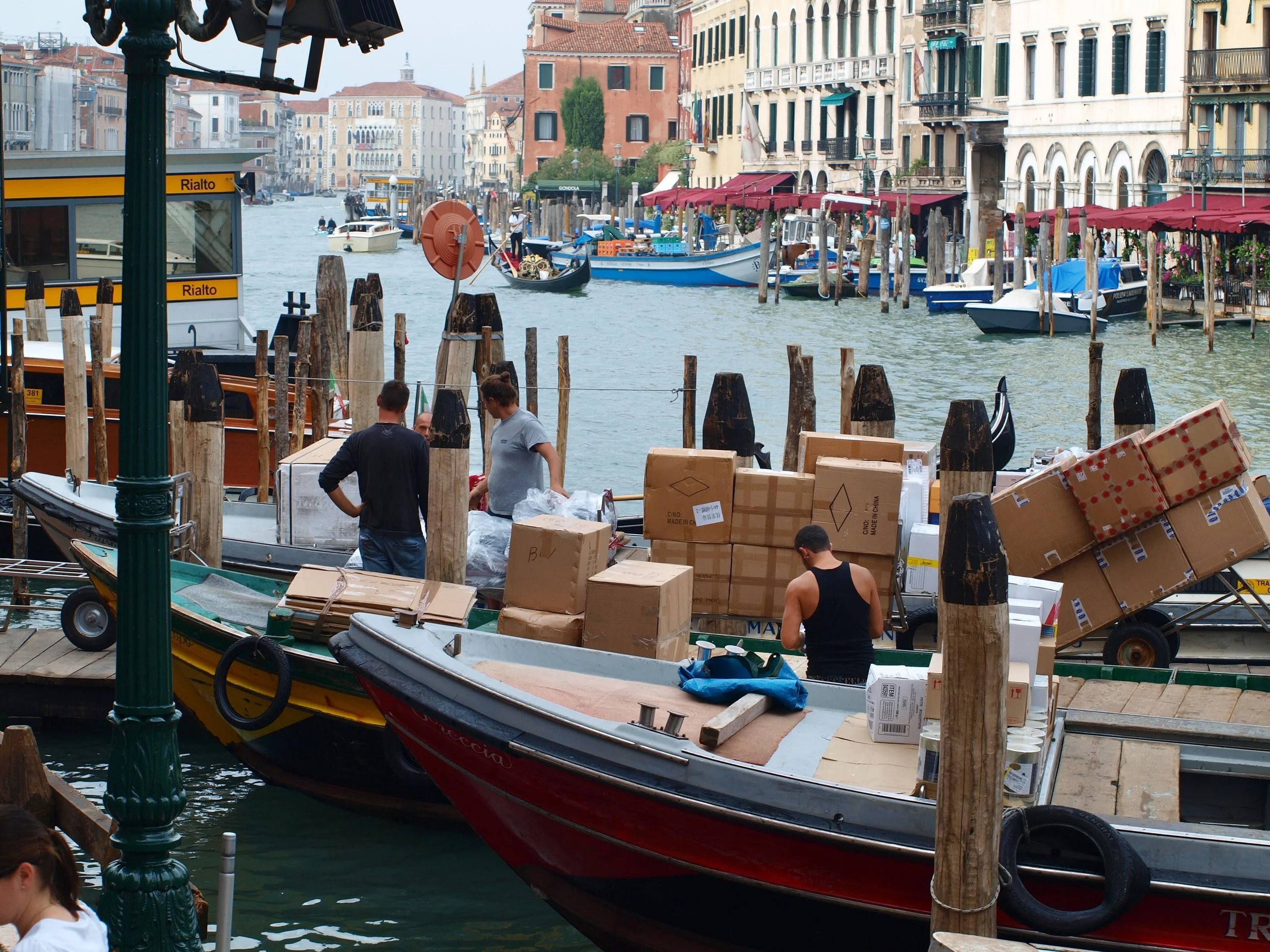 Une scène de canaux à Venise avec des bateaux, des bâtiments colorés, des tables de restaurant sous des auvents rouges, et des personnes travaillant ou se préparant à charger des colis dans un bateau.