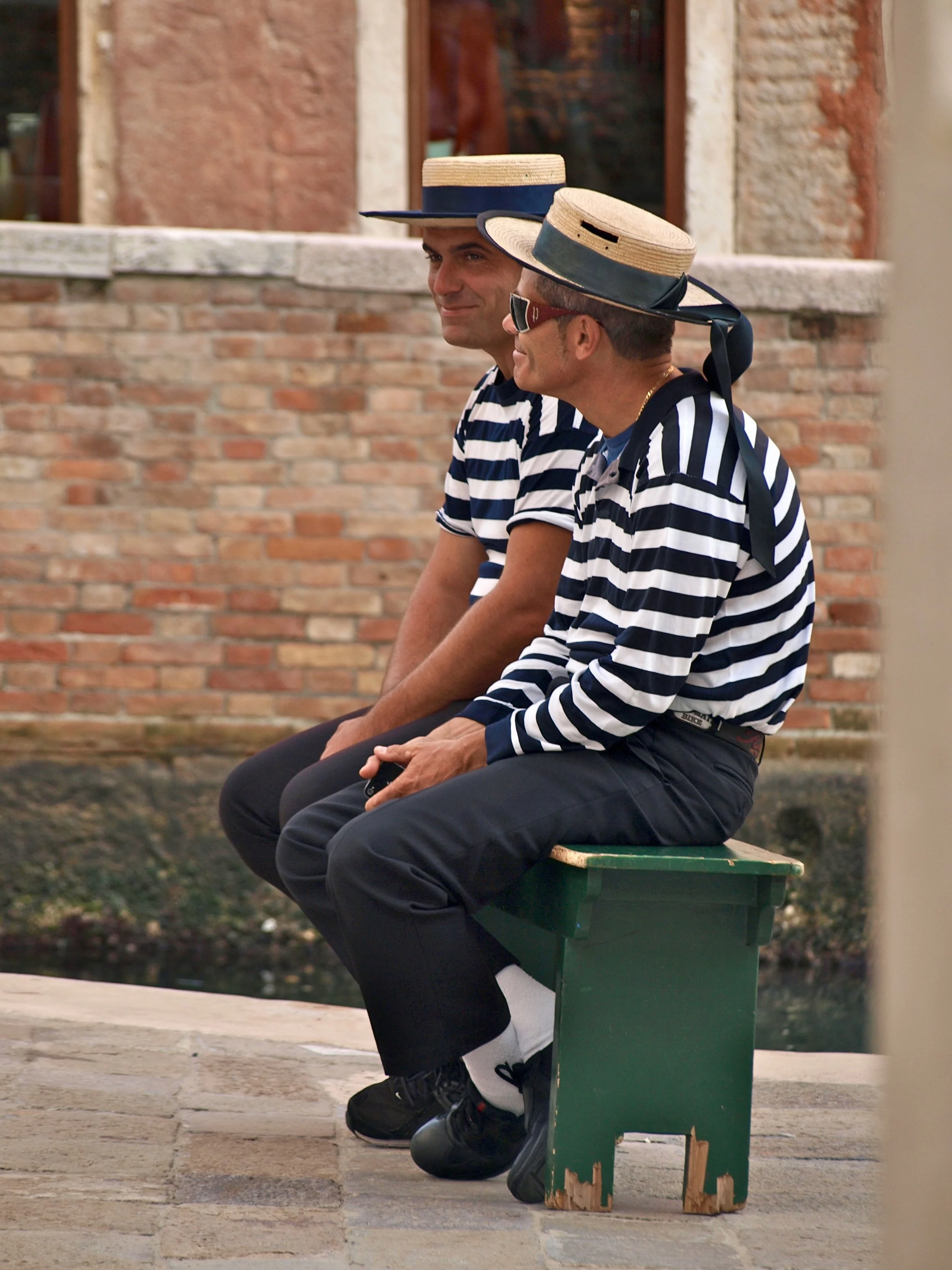 Deux hommes assis sur un banc en bois, portant des chapeaux de paille à rayures bleues et blanches, des lunettes de soleil, et des vêtements à rayures, devant un mur de briques.