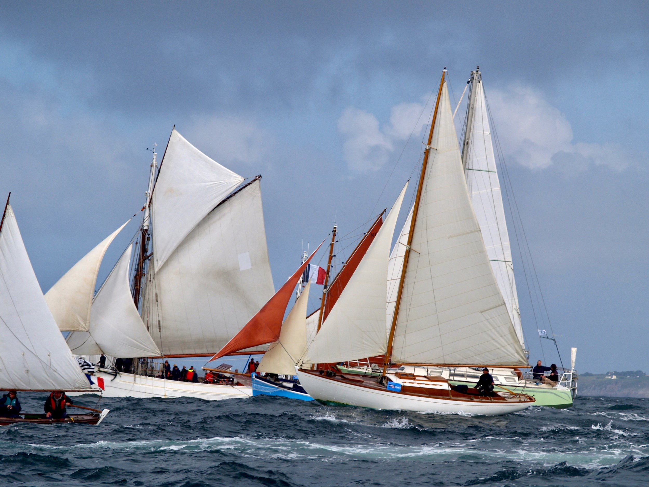 Plusieurs voiliers de course avec voiles blanches et brunes à la mer lors d'une régate, ciel couvert, vue de côté.