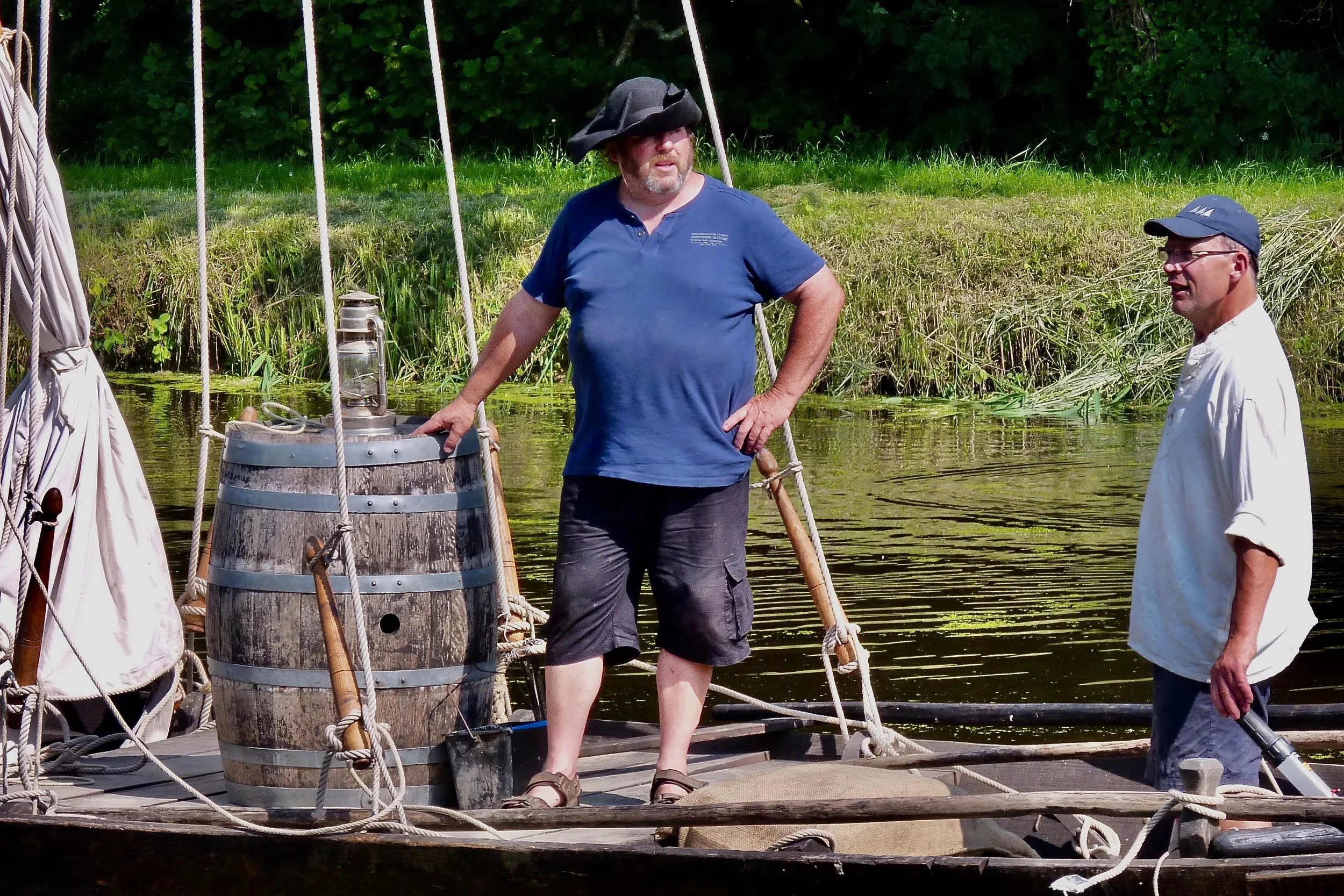 Deux hommes debout sur une barque en bois sur l'eau, entourés d'arbres et de végétation, portant des vêtements décontractés et chapeaux, avec des cordages et un tonneau en bois sur la barque.