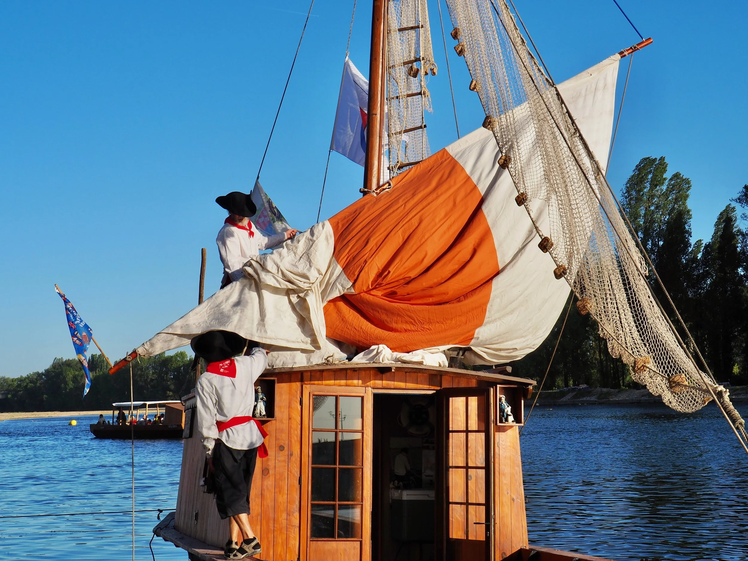 Une maquette de bateau en bois avec des voiles en tissu orange et blanc flotte sur l'eau, un homme en costume traditionnel avec un chapeau noir et un foulard rouge ajuste la voile. Le fond montre un ciel bleu clair et des arbres au loin.