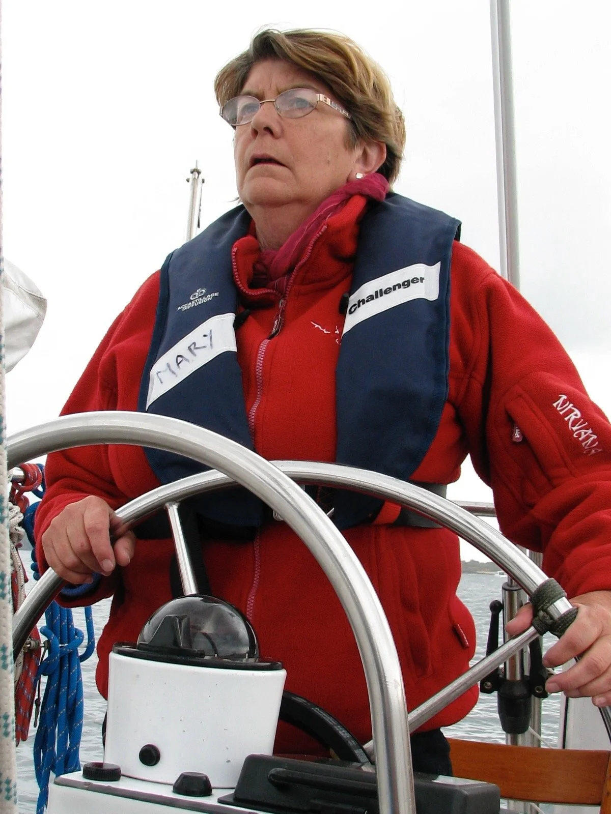 Une femme âgée, portant une veste rouge et un gilet pare-balles, se tient au gouvernail d'un bateau, avec une étiquette portant le nom "Mary".