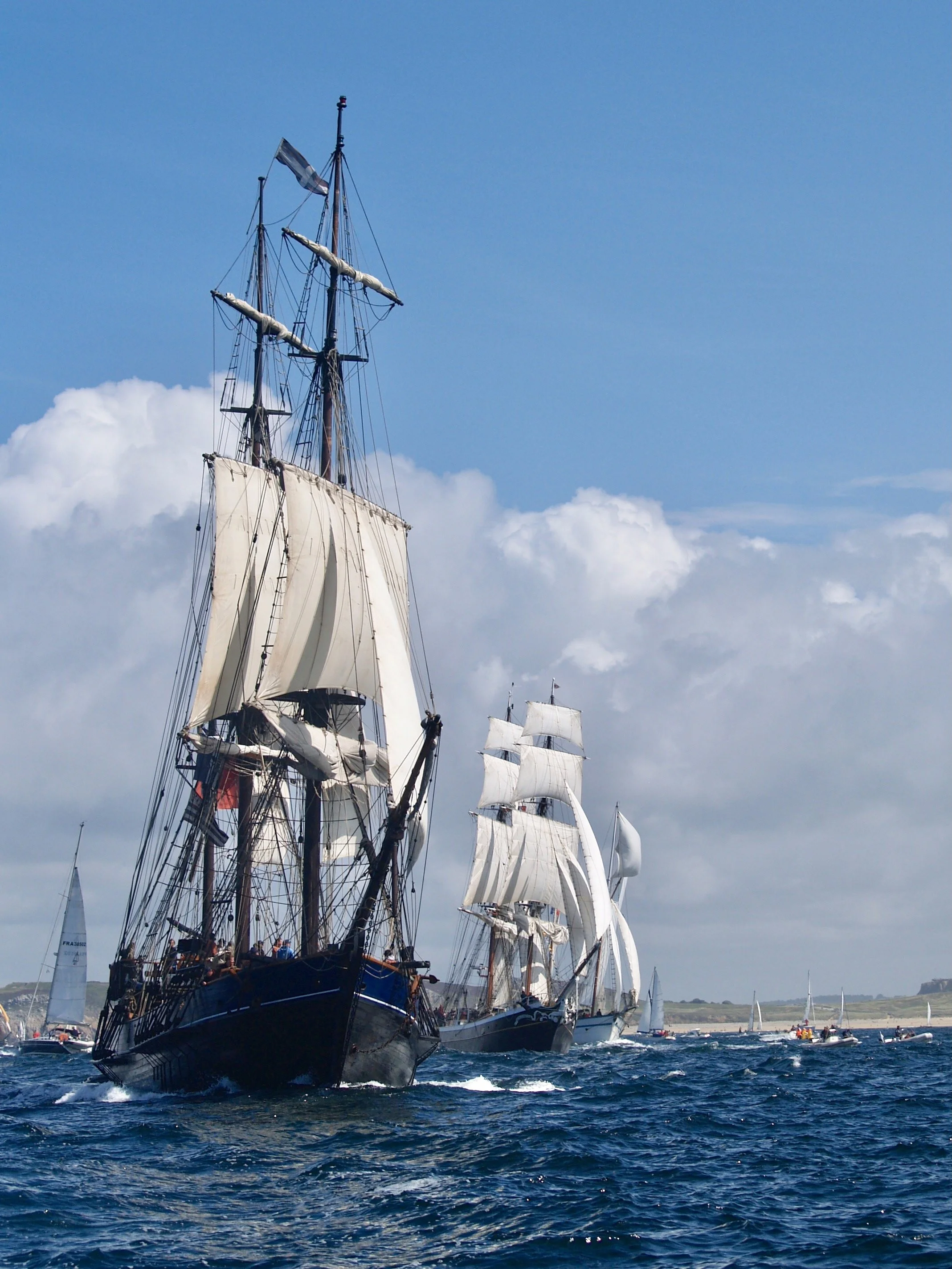 Un voilier traditionnel à plusieurs mâts avec des voiles blanches déployées, naviguant sur une mer calme avec un ciel partiellement nuageux.