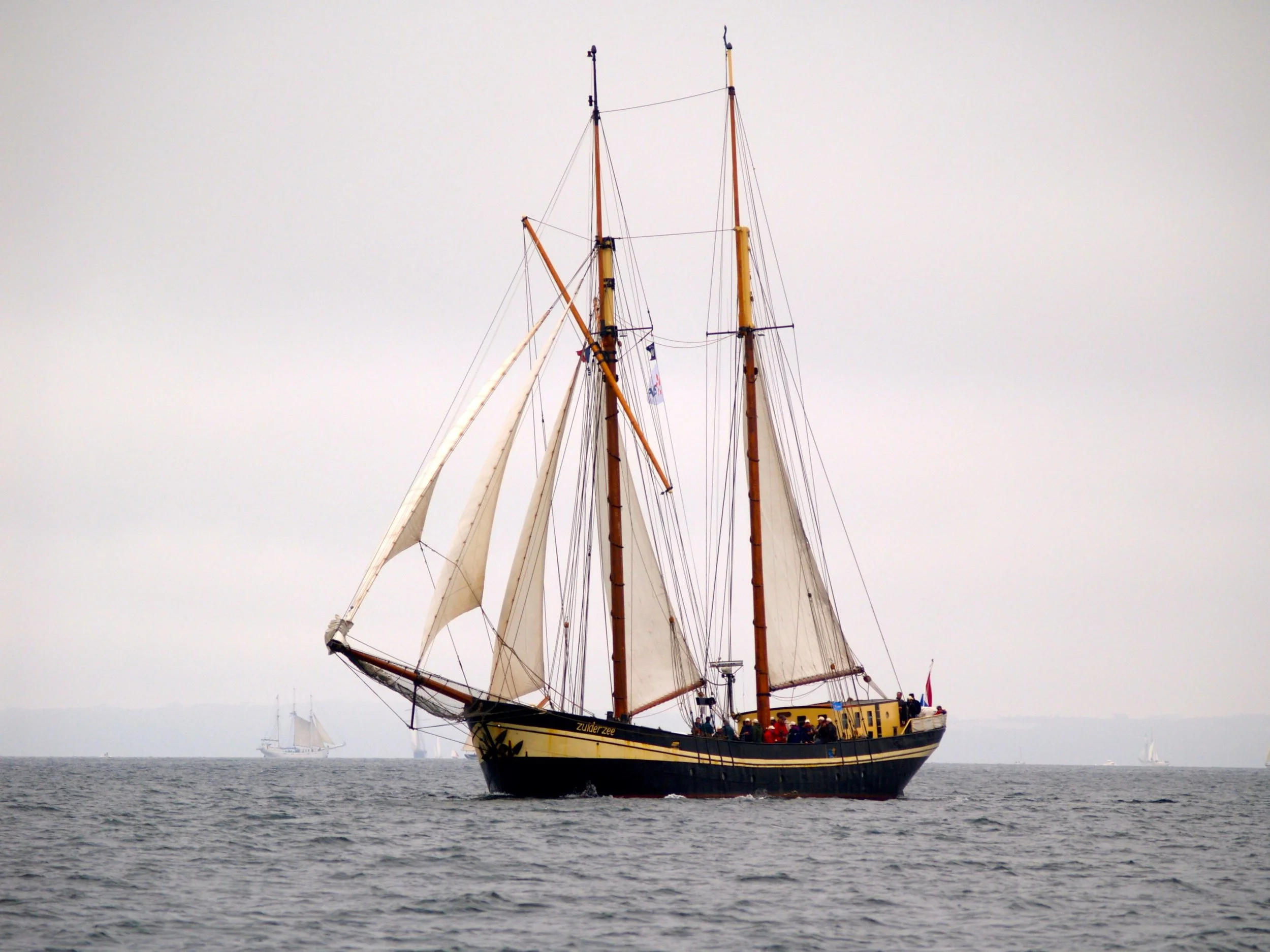 Un grand voilier avec des voiles en toile beige et des mâts en bois navigue sur la mer. La mer est calme et le ciel est gris. Il y a d'autres voiliers à l'arrière-plan.