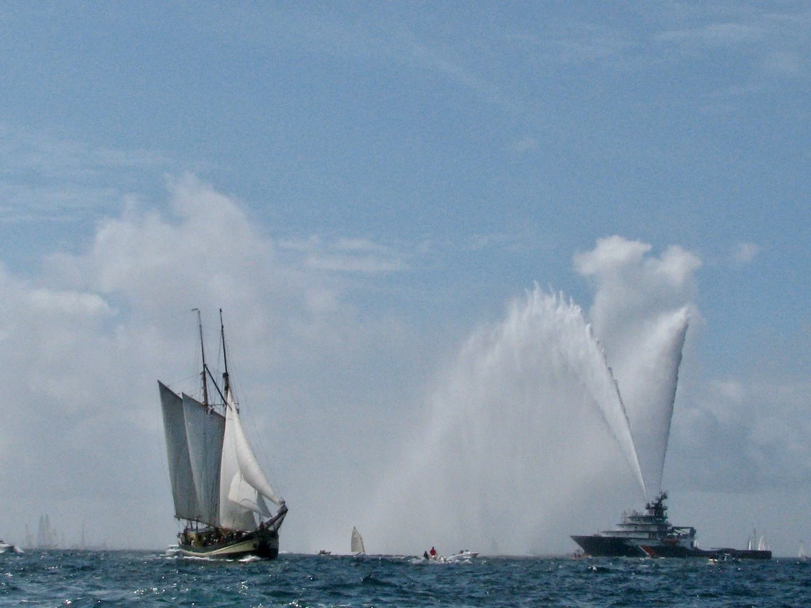 Deux grands bateaux en mer, un avec des voiles traditionnels et l'autre un yacht moderne. Les bateaux crachent de l'eau en l'air, sous un ciel nuageux.