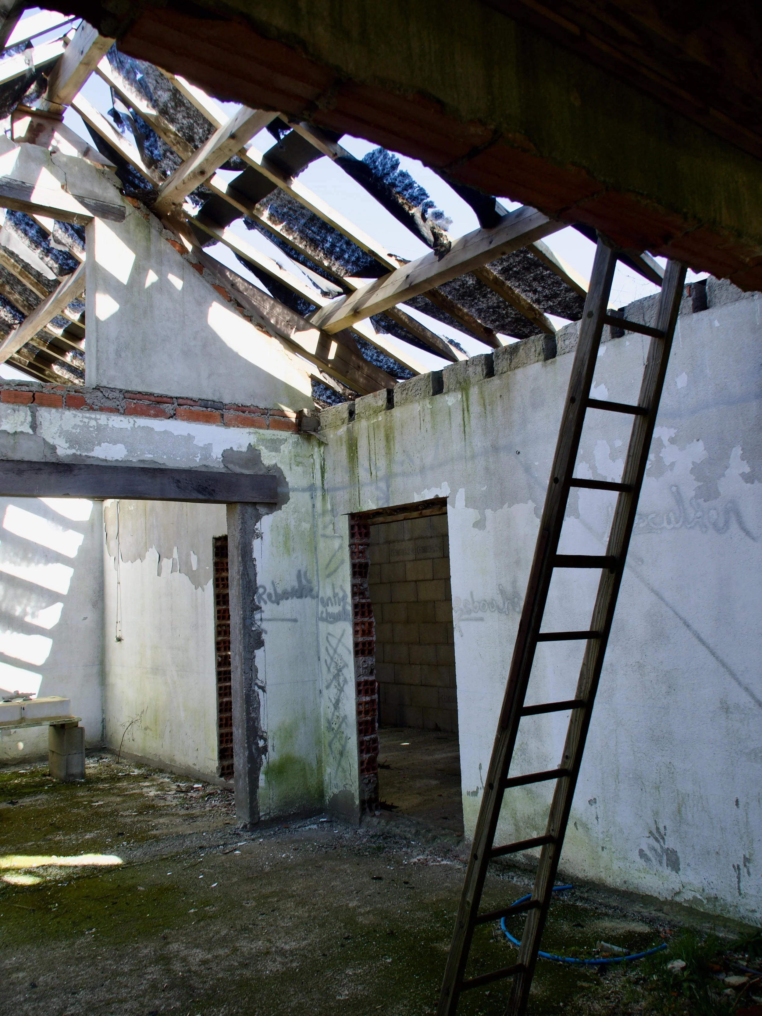 Une pièce en construction avec un plafond inachevé, des murs en béton et briques, un escalier en bois, et des murs peints en blanc avec des traces de graffiti.