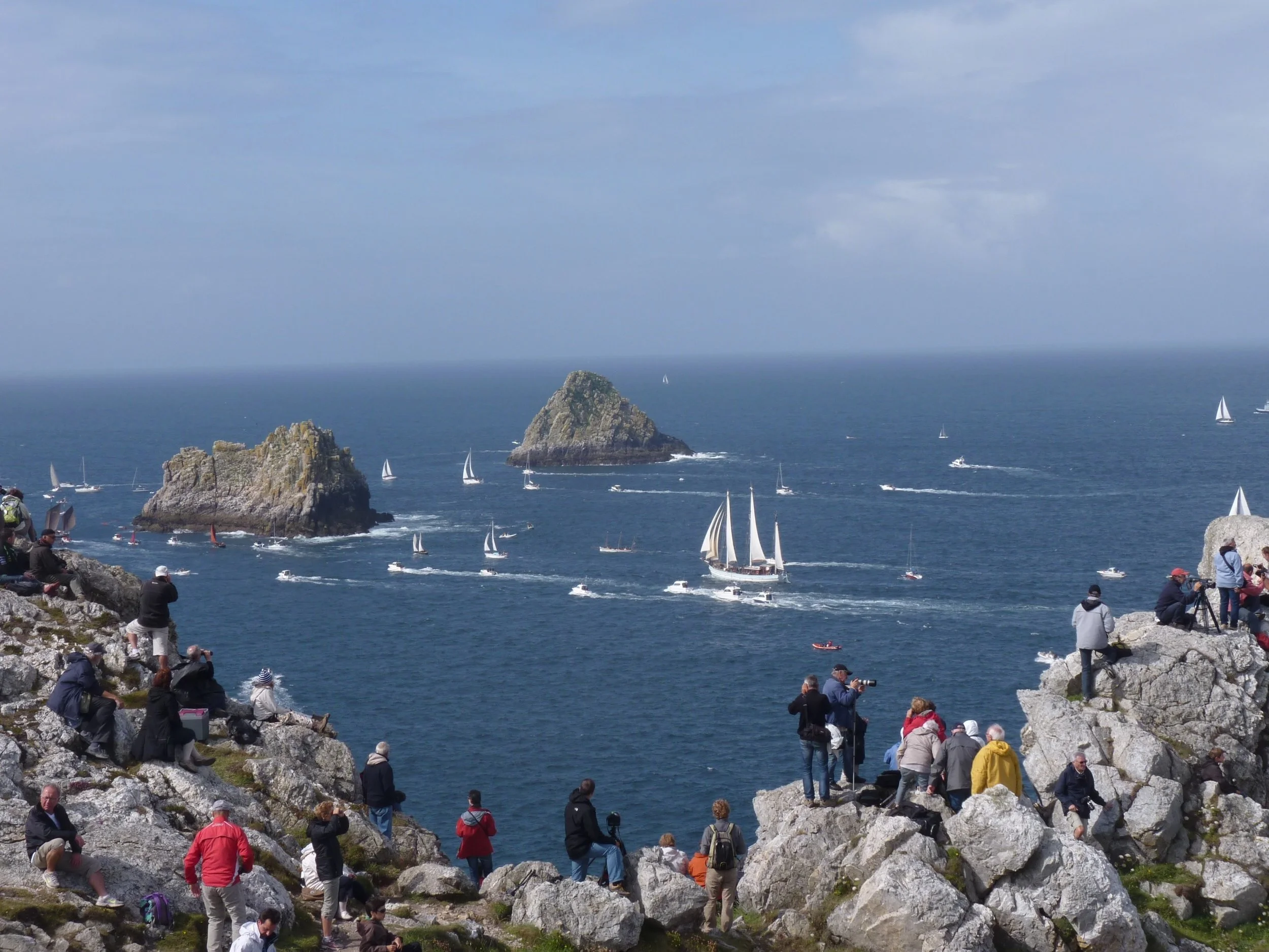 Une foule de personnes assises et debout sur des rochers surplombant la mer, avec des voiliers naviguant au large d'une île rocheuse dans un ciel partiellement nuageux.
