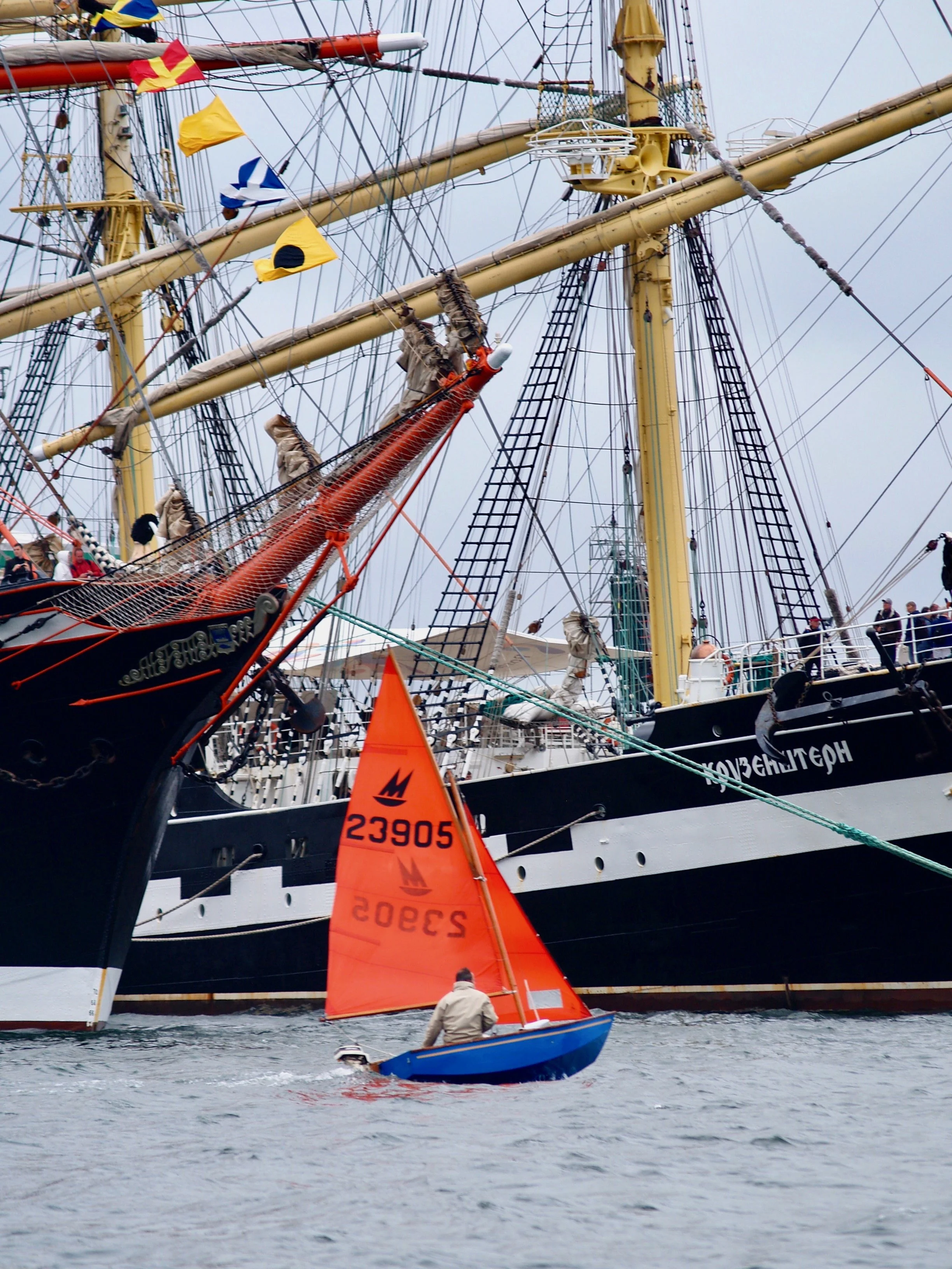 Un voilier de grande taille avec des voiles colorées en arrière-plan, et une petite embarcation avec une voile orange en avant, sur l'eau.