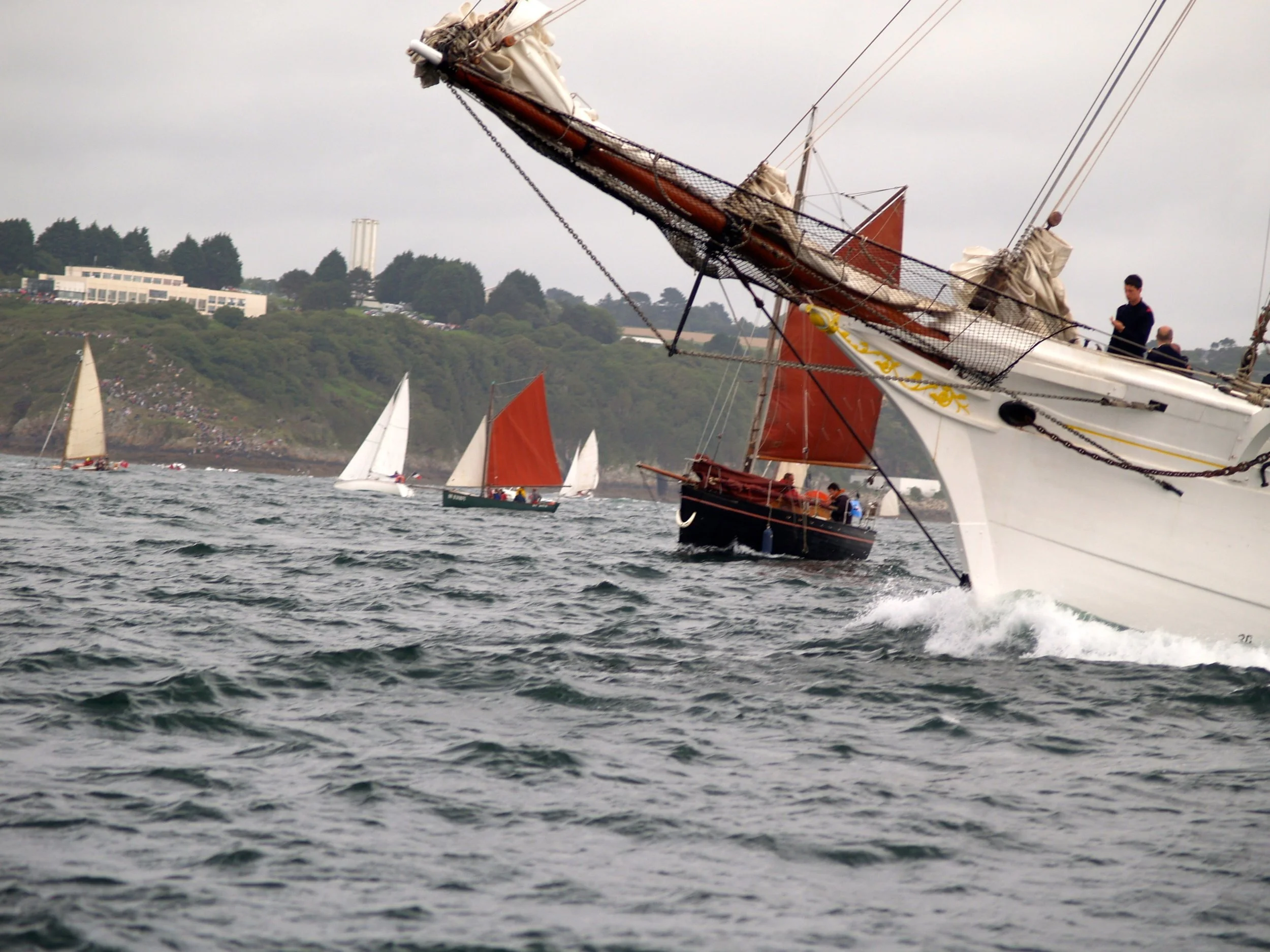 Navigation de bateaux à voile lors d'une régate en mer, avec plusieurs voiliers en compétition et un paysage côtier en arrière-plan.