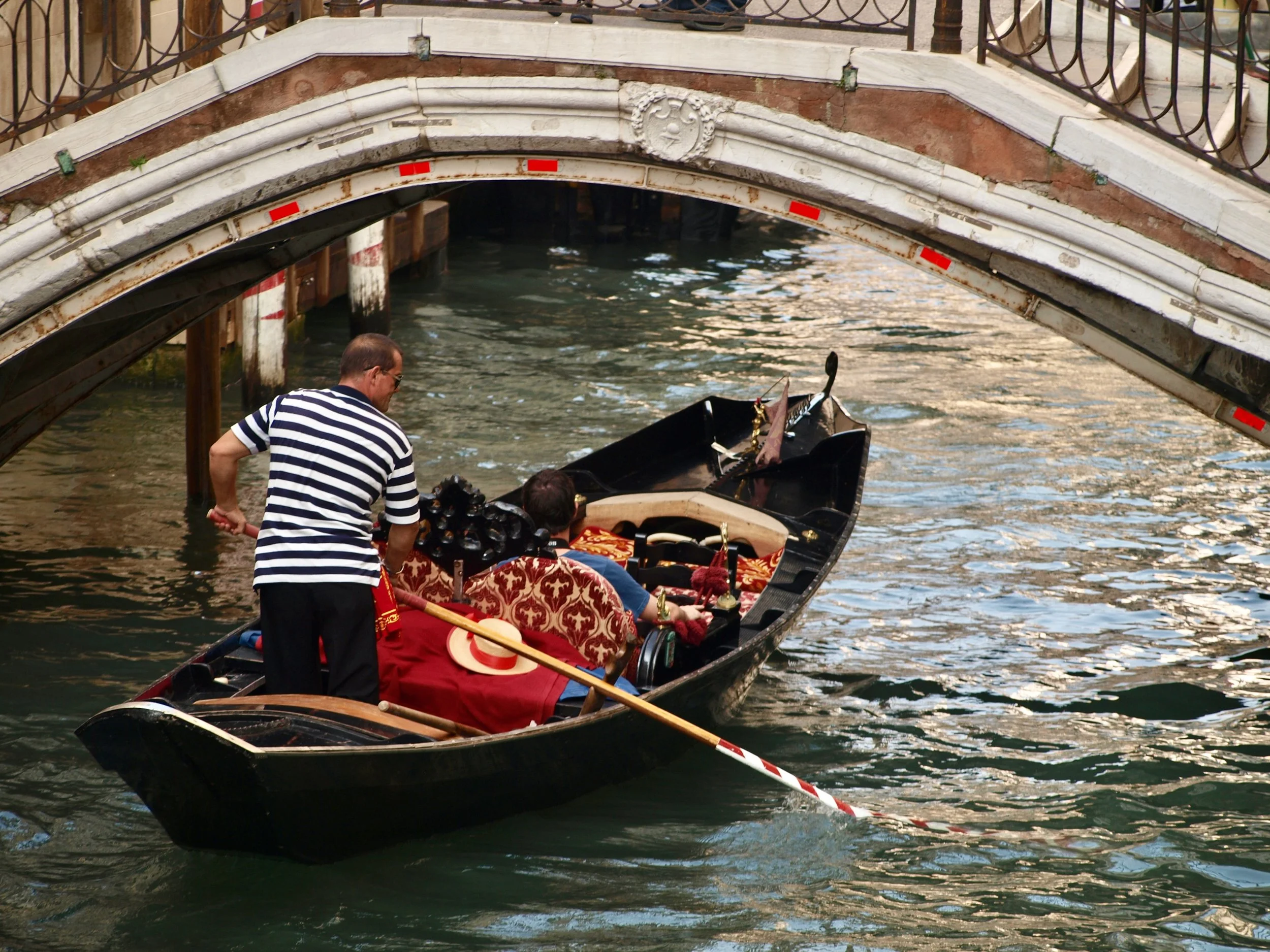 Gondolier en striped shirt manoeuvrant une gondole devant un pont en pierre orné de détails sculptés, dans une canal de Venise.