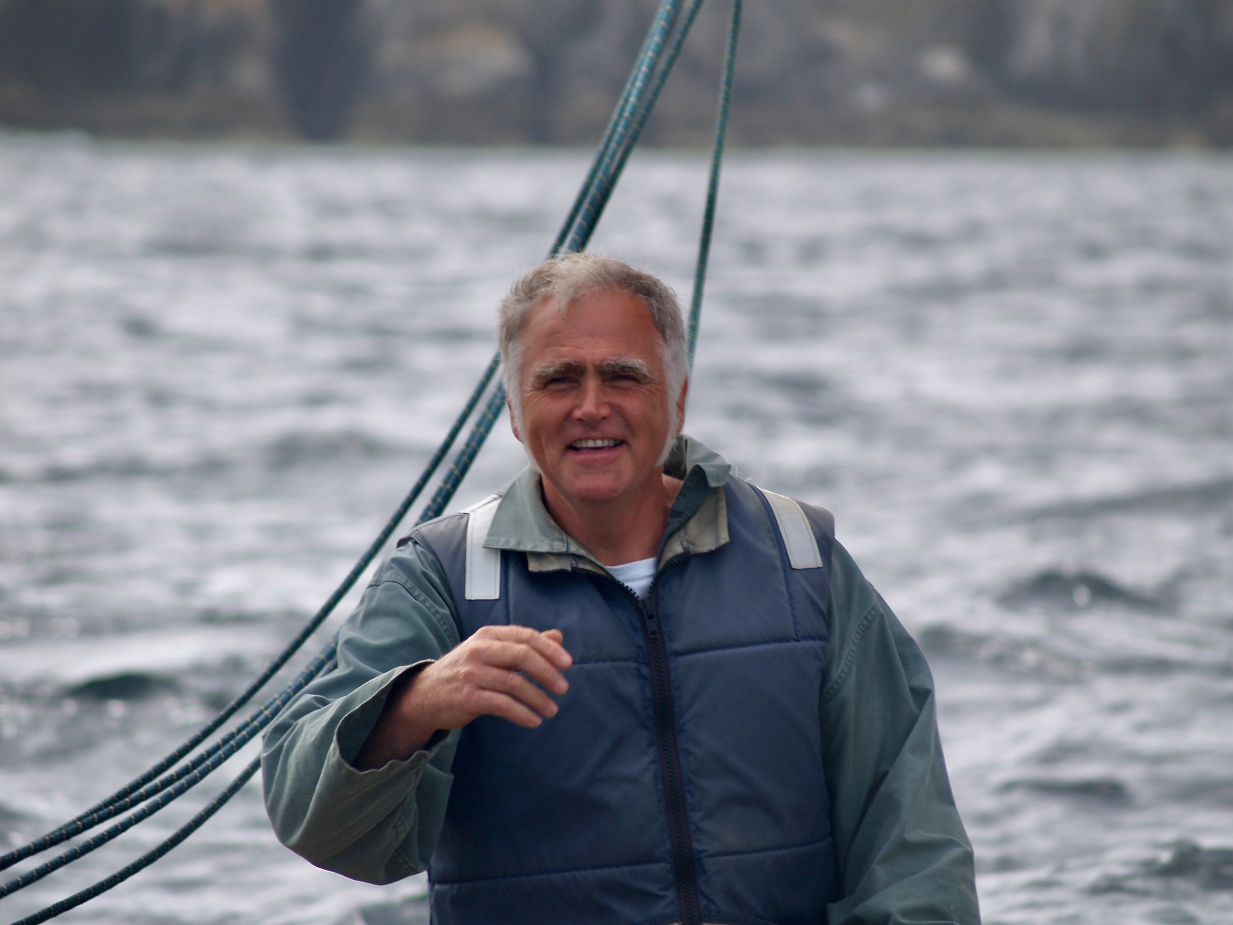 Un homme âgé souriant à la statue de la liberté, portant une veste grise et un sac à dos, sur un bateau en mer.