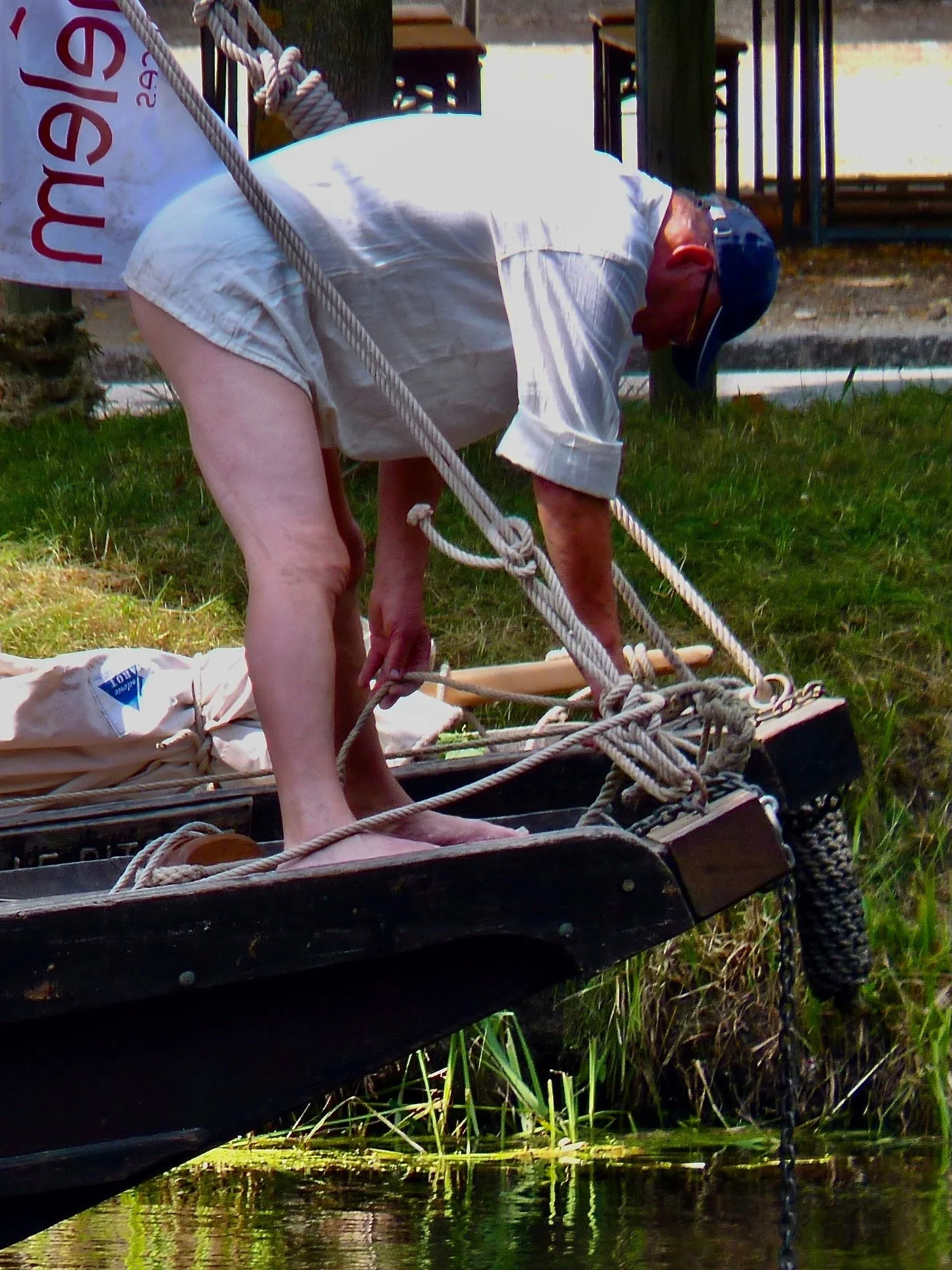Une personne en maillot de bain, portant un chapeau de vélo, penchée vers une barque en bois dans l'eau, avec des cordages et un sac blanc sur la barque.